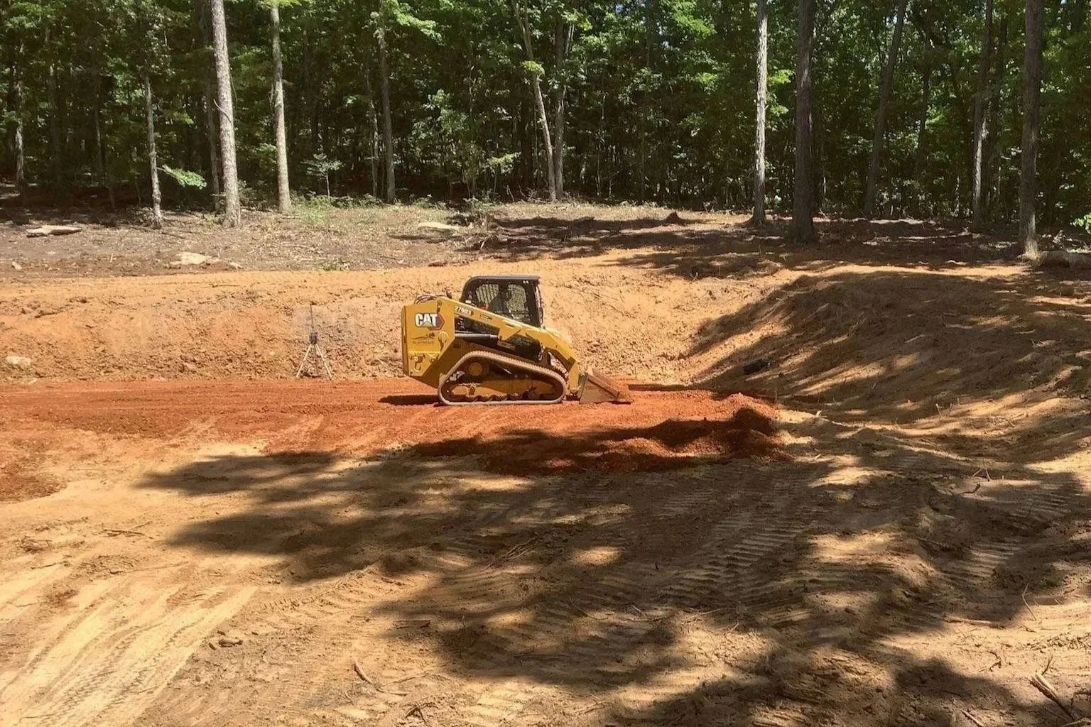 A yellow Caterpillar compact track loader working on a construction site with cleared dirt, surrounded by trees in a forested area.