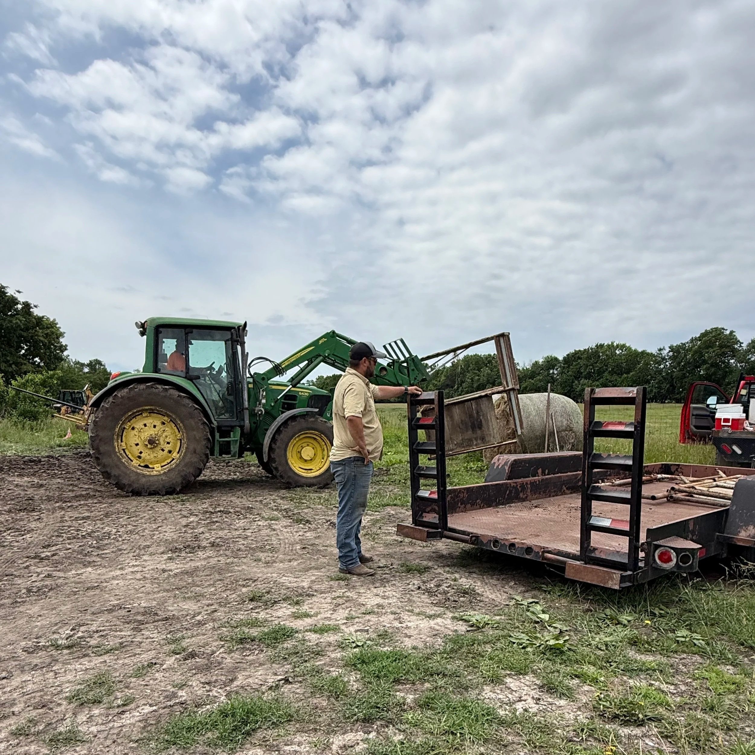 A man and a woman at a farm, with a tractor and trailer, and fields in the background under a cloudy sky.