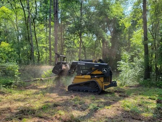 A small John Deere compact track loader operating in a wooded area, clearing or moving dirt among trees.