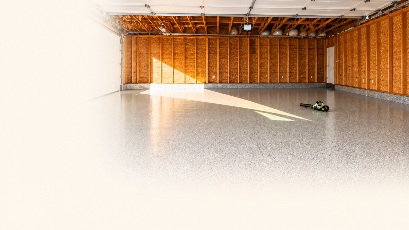 Empty garage with unfinished wood walls, concrete floor, and a partially open overhead door. A leaf blower tool is on the floor near the right side.