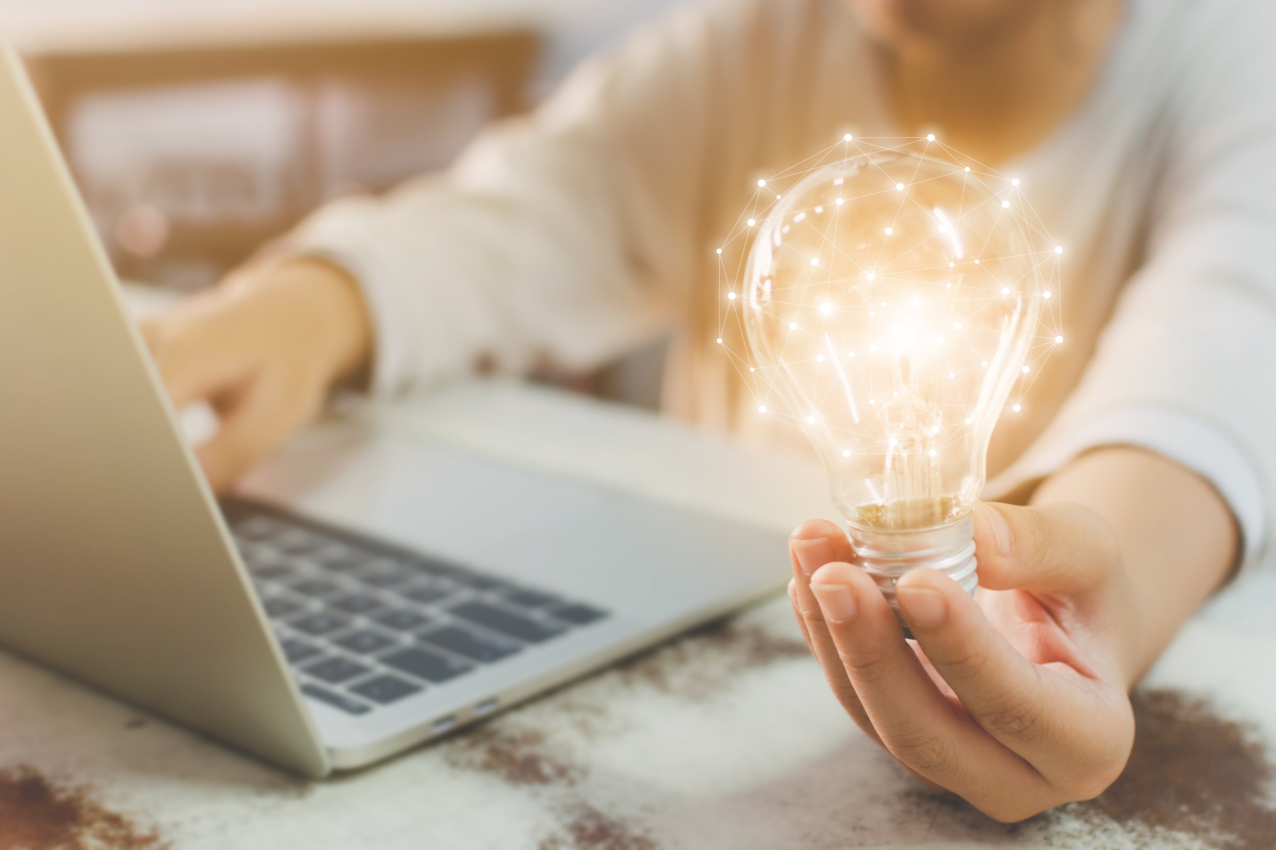 Person holding a light bulb that has digital connections and glowing points around it, with a laptop in the background.