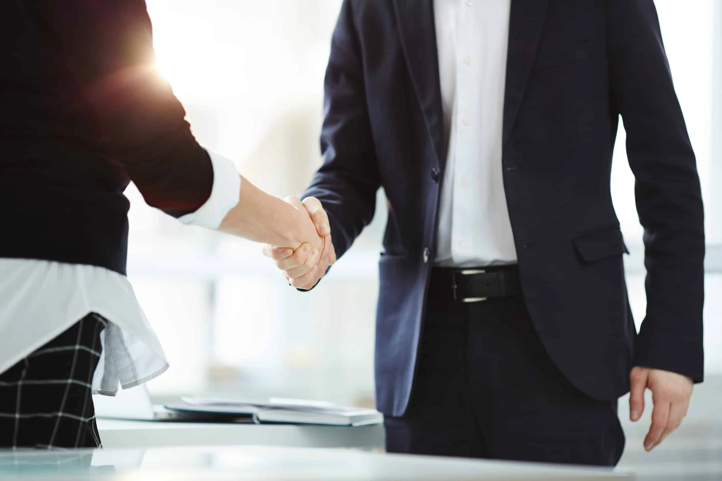 Two people shaking hands in a professional setting, with a focus on the handshake and partial view of their bodies, one wearing a business suit.