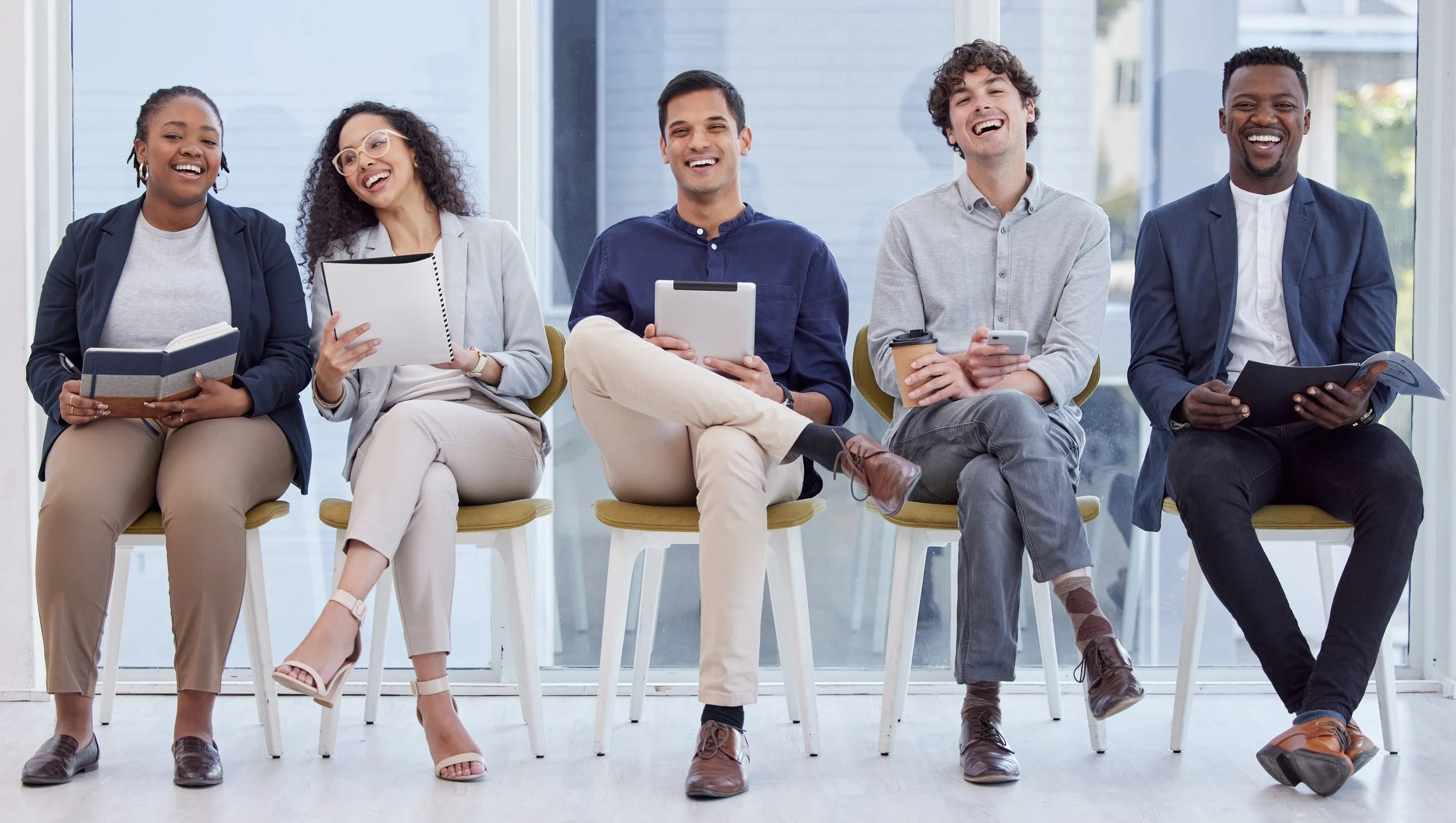 Diverse group of five young adults sitting in chairs, laughing and smiling in a modern indoor space with large windows.