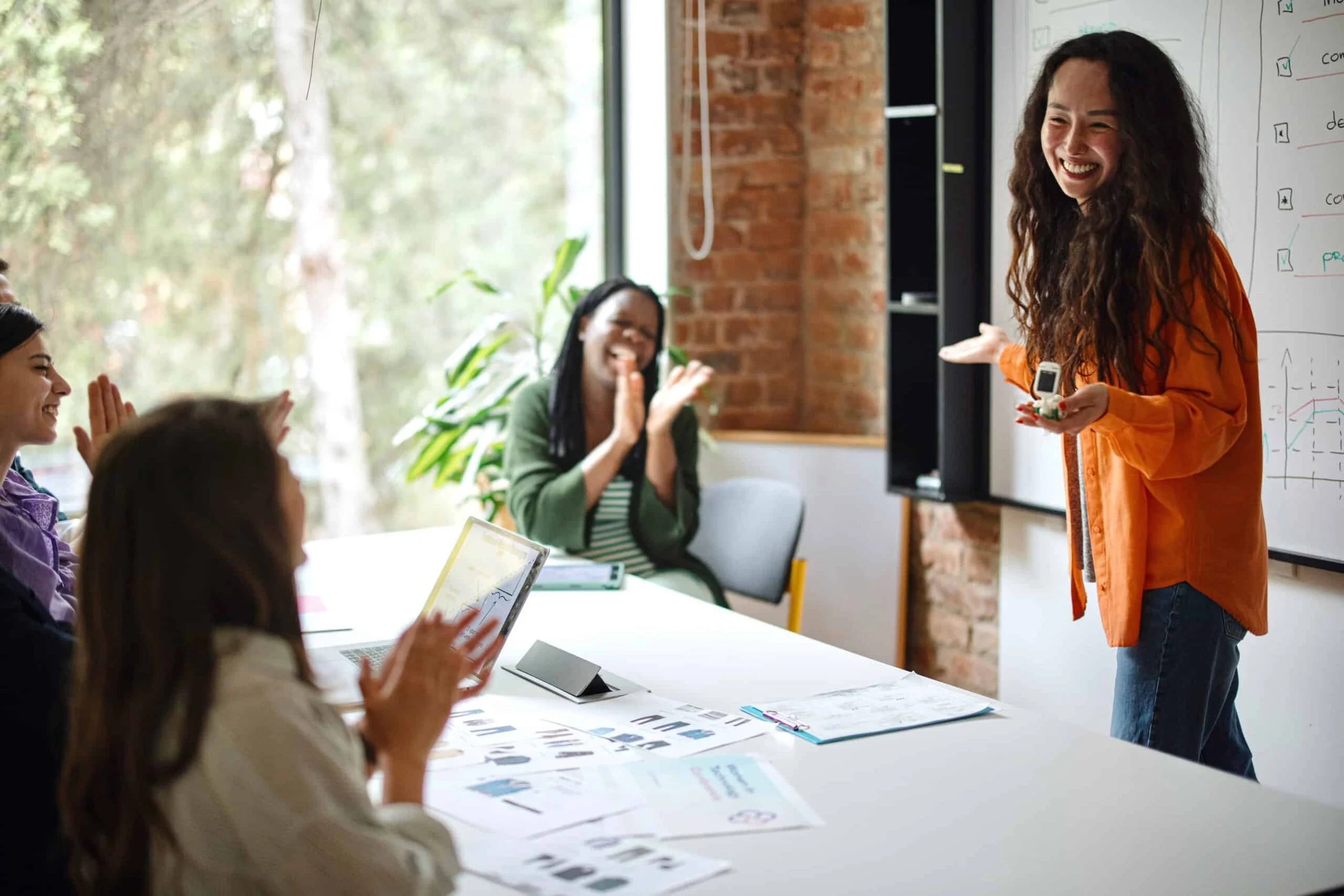 A woman with curly hair and a bright orange shirt smiling and presenting in front of a whiteboard with charts and checklists, while a group of women, some with laptops, applaud in a modern, well-lit room with large windows and brick walls.