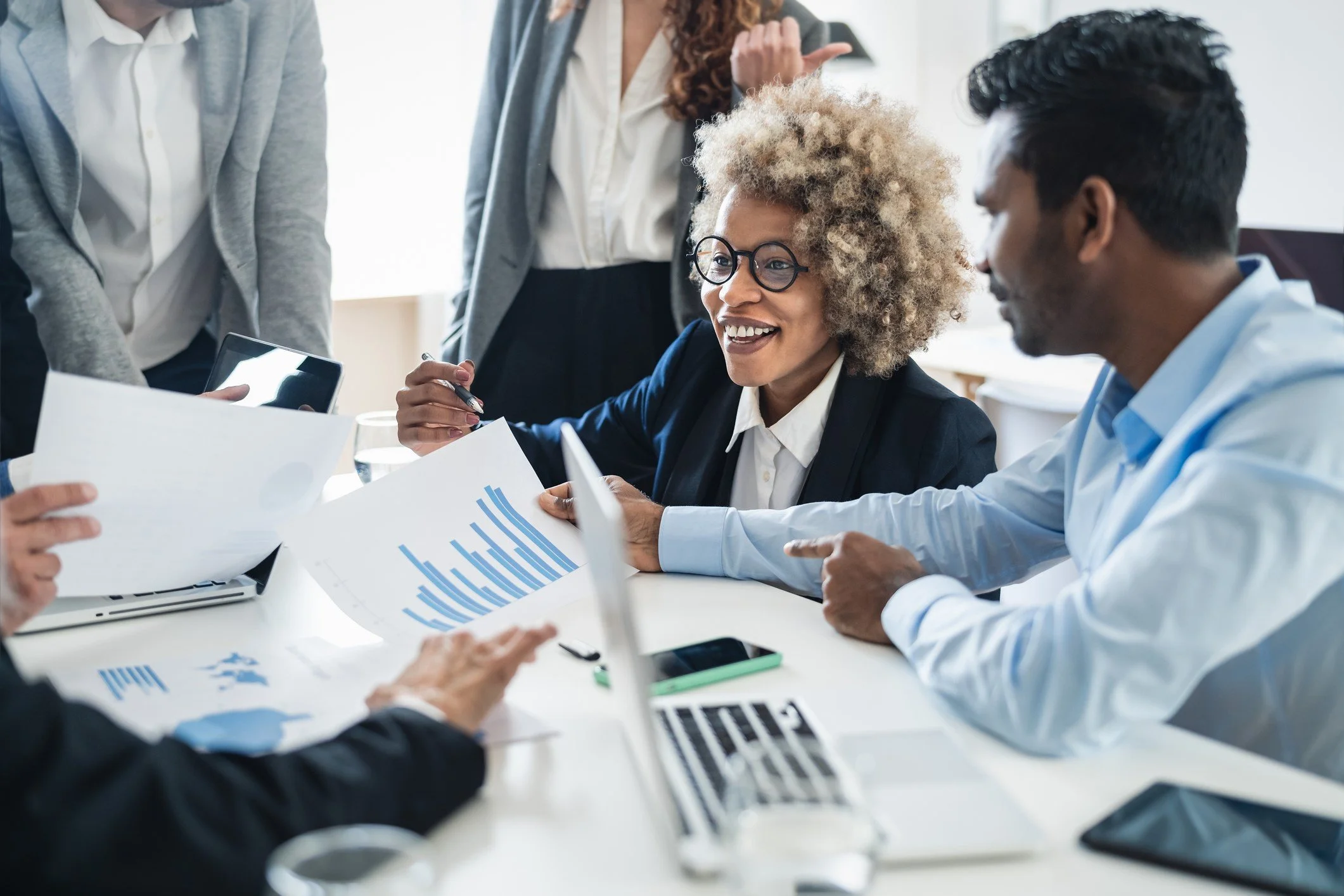 Group of diverse professionals having a discussion in a modern office with documents, charts, and a laptop on the table.