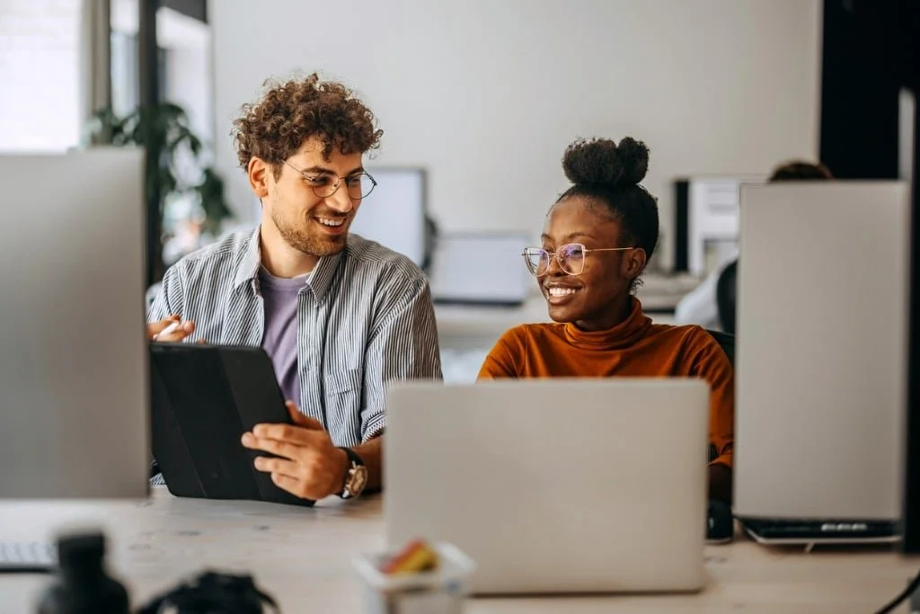 Two colleagues, a man with curly hair and glasses and a woman with glasses and a bun, are smiling and talking in an office surrounded by computer monitors, with one holding a tablet.