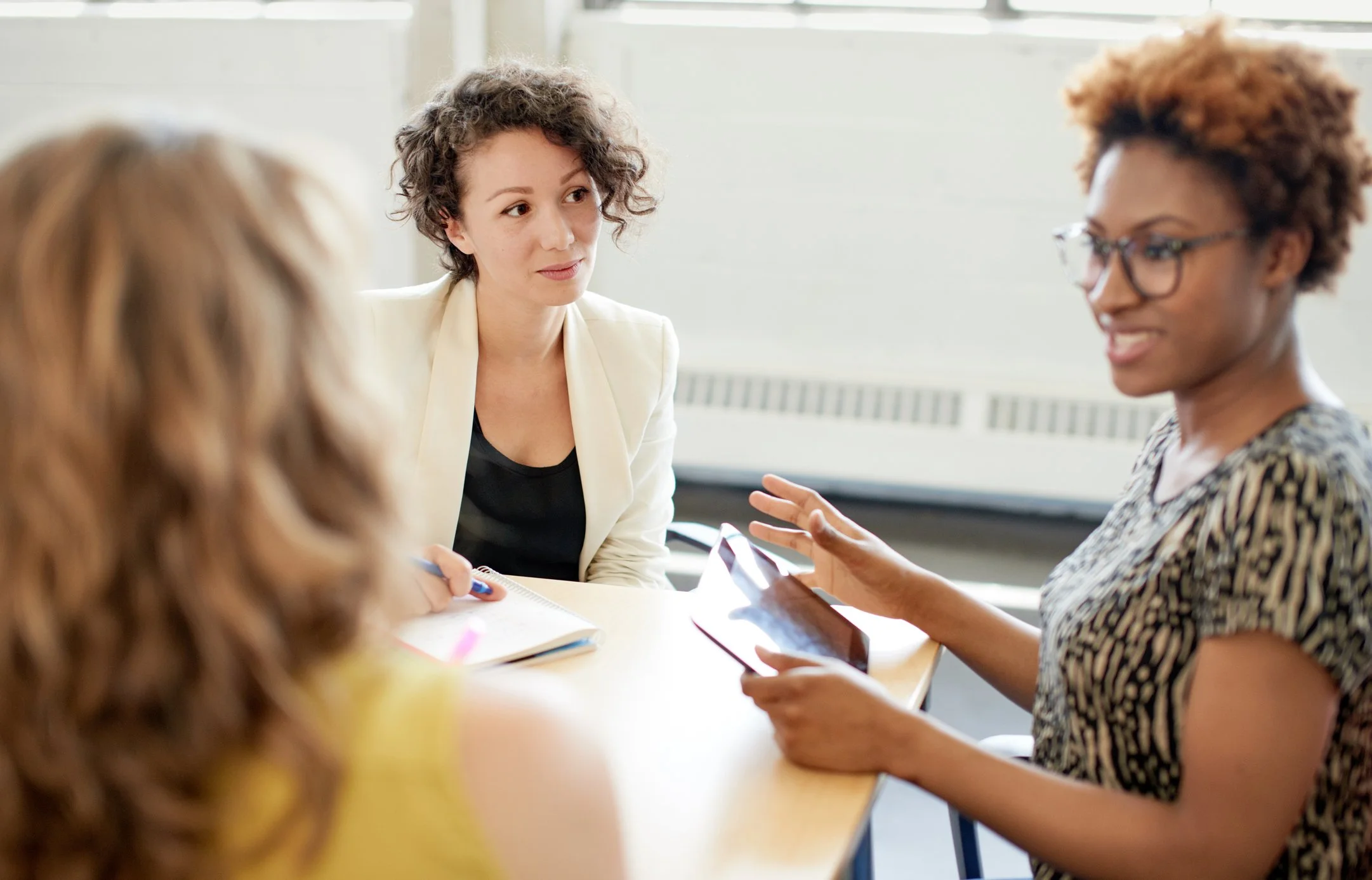 Four women engaged in a discussion at a table in a bright office, with notebooks and a tablet in front of them.