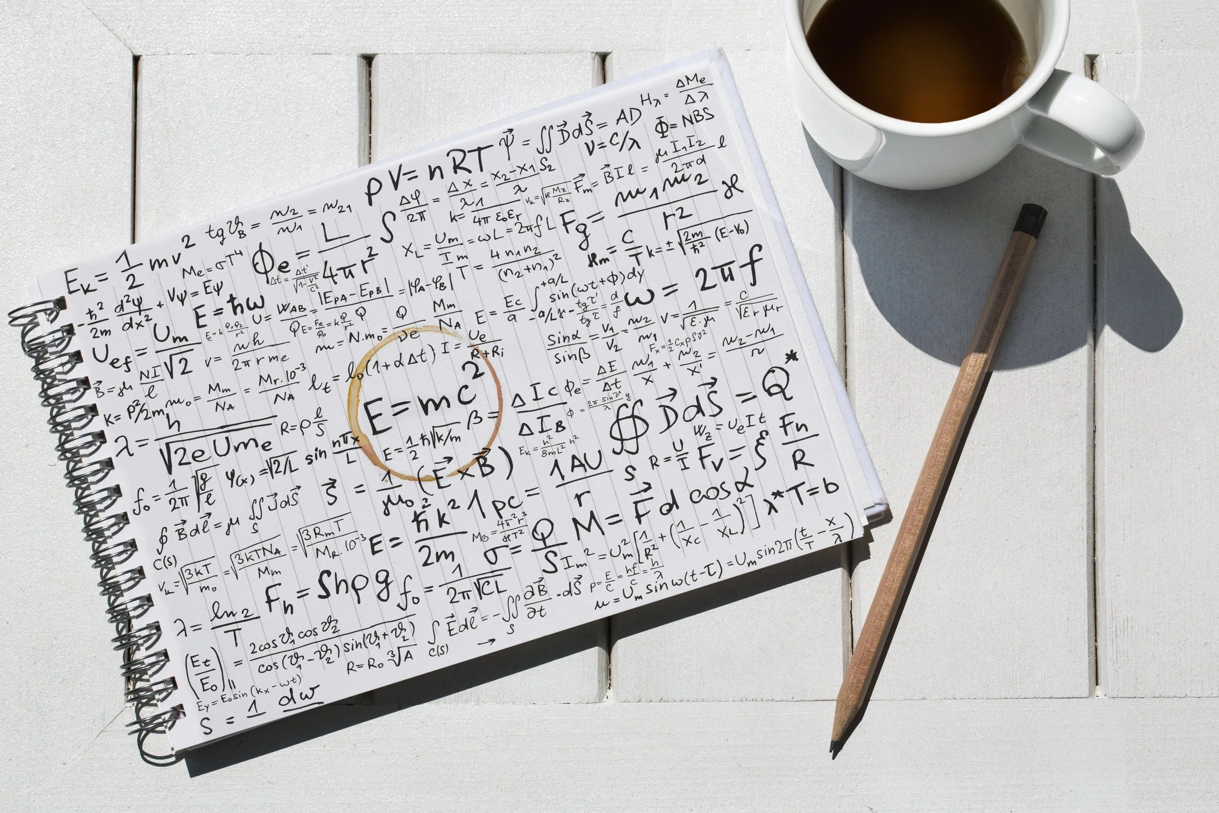 A notebook with pages filled with handwritten physics and math equations, a cup of coffee, a pen, and a shadow on a white surface.