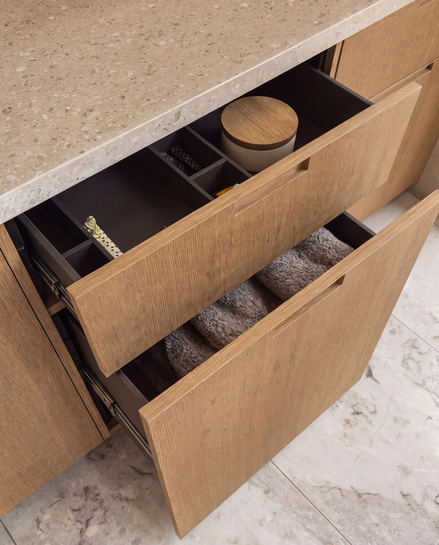 Open wooden kitchen drawer containing a ceramic container with a wooden lid, miscellaneous small items, and a cloth underneath the drawer, with marble tile flooring visible.