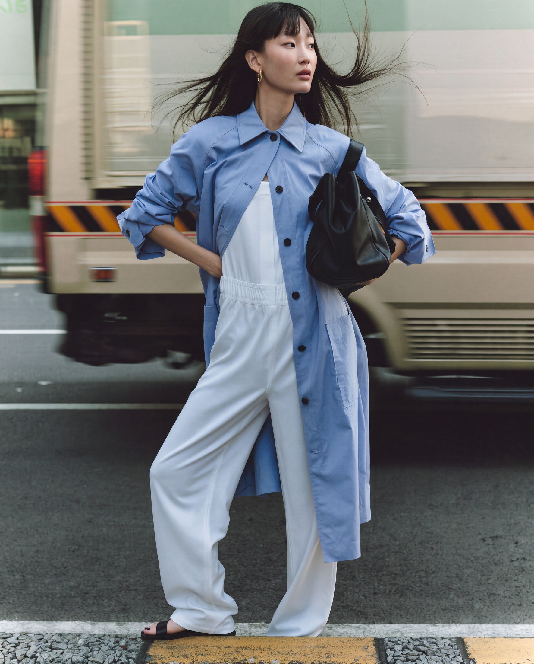 A woman with long dark hair and earrings standing in front of moving buses in an urban setting, wearing a blue button-up shirt over a white dress and white wide-leg pants, with her hand on her hip.