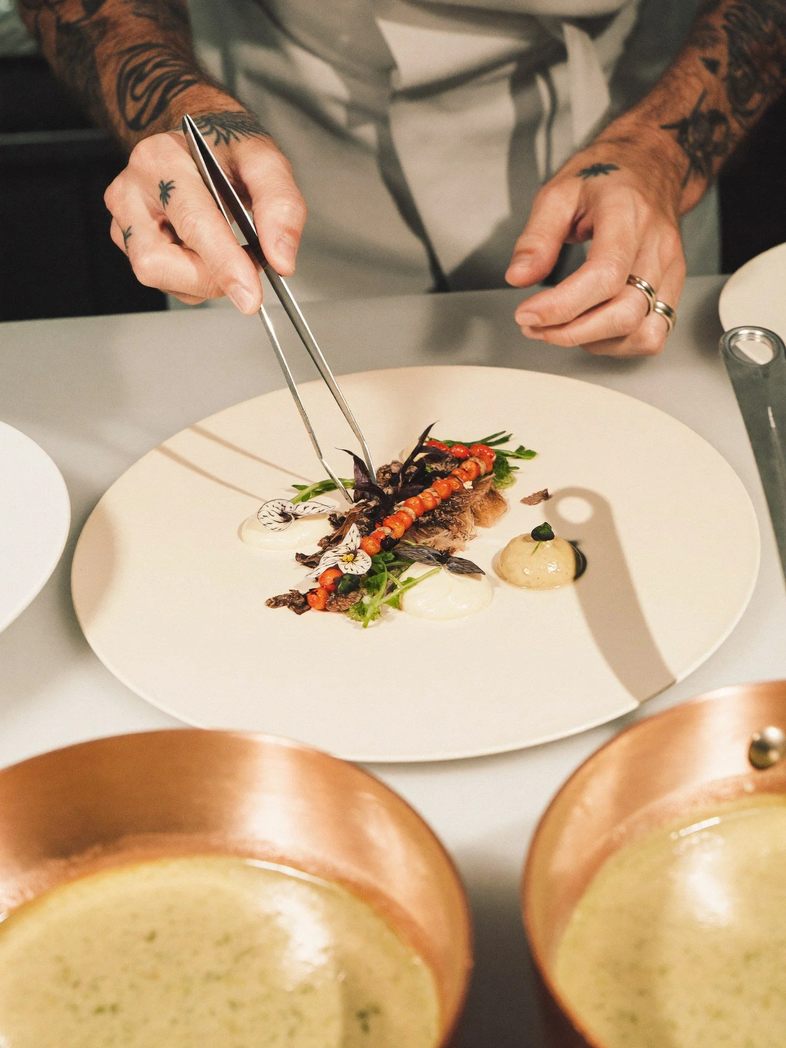 A chef prepares a dish with a fish fillet topped with vegetables and edible flowers, garnished with sauce, while using tongs for plating.