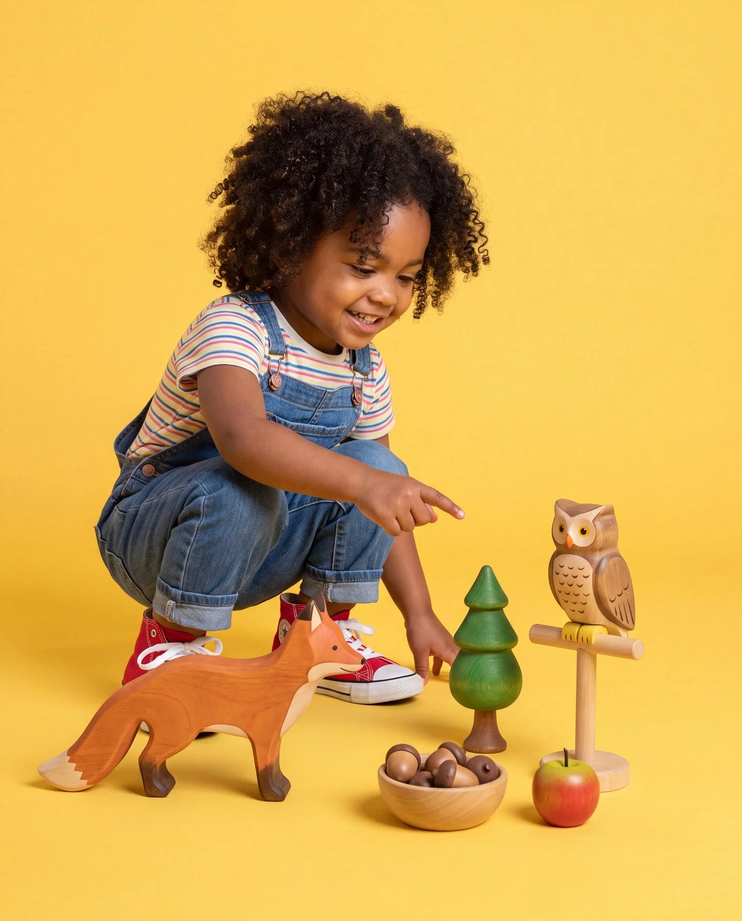 A young girl with curly hair crouches down, smiling, and playing with wooden animal toys, trees, and an apple against a yellow background.