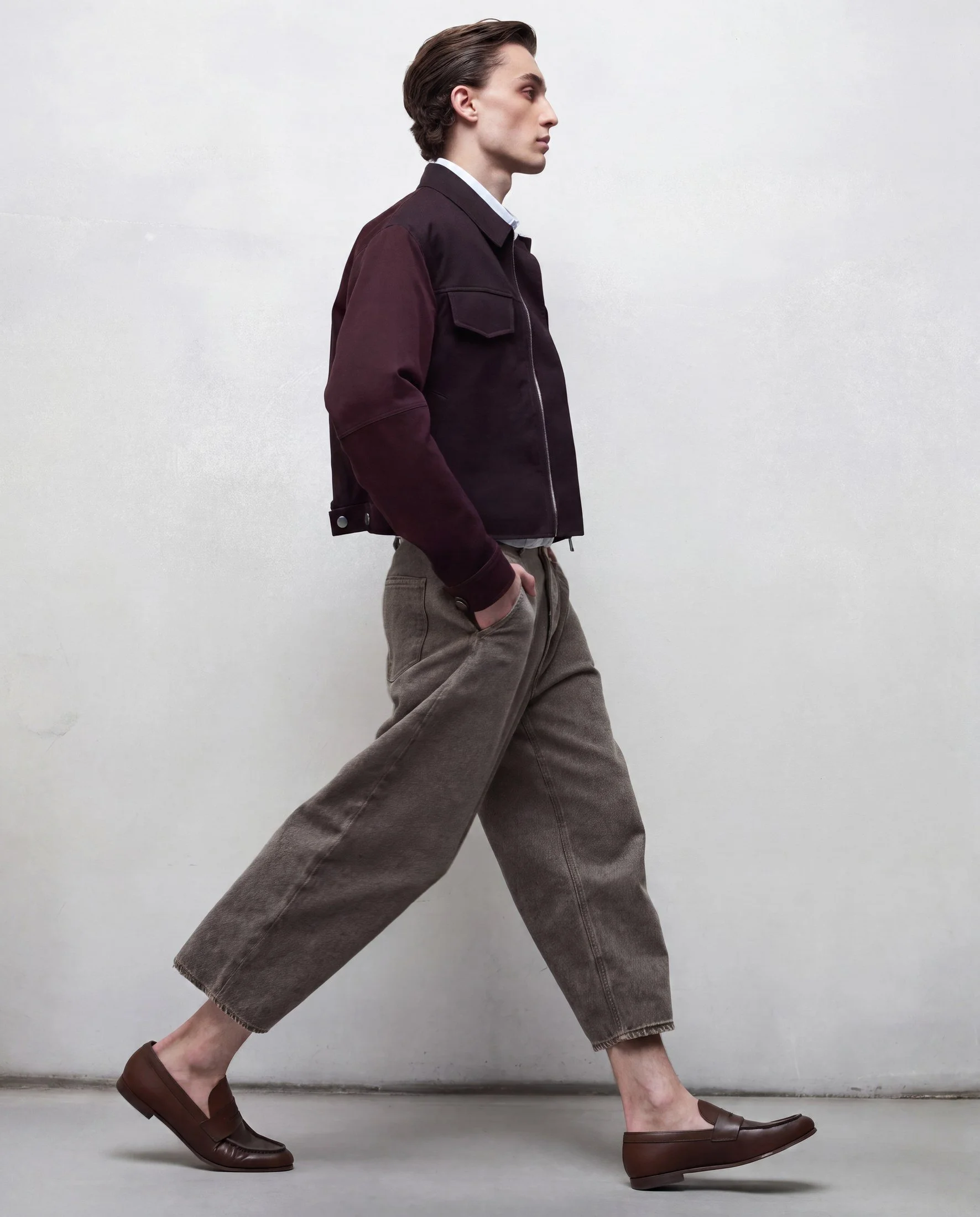 Person with brown slip-on shoes, loose beige pants, a dark jacket, white shirt, standing sideways against a plain white wall.