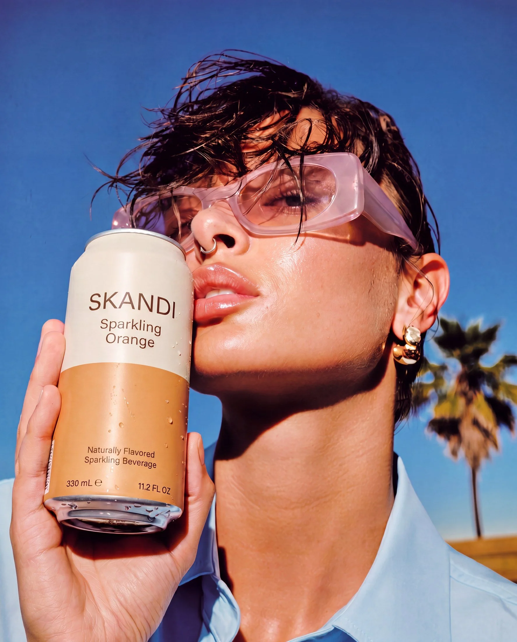 A person with wet hair and sunglasses holding a can of SKANDI Sparkling Orange in front of their face, against a blue sky background.
