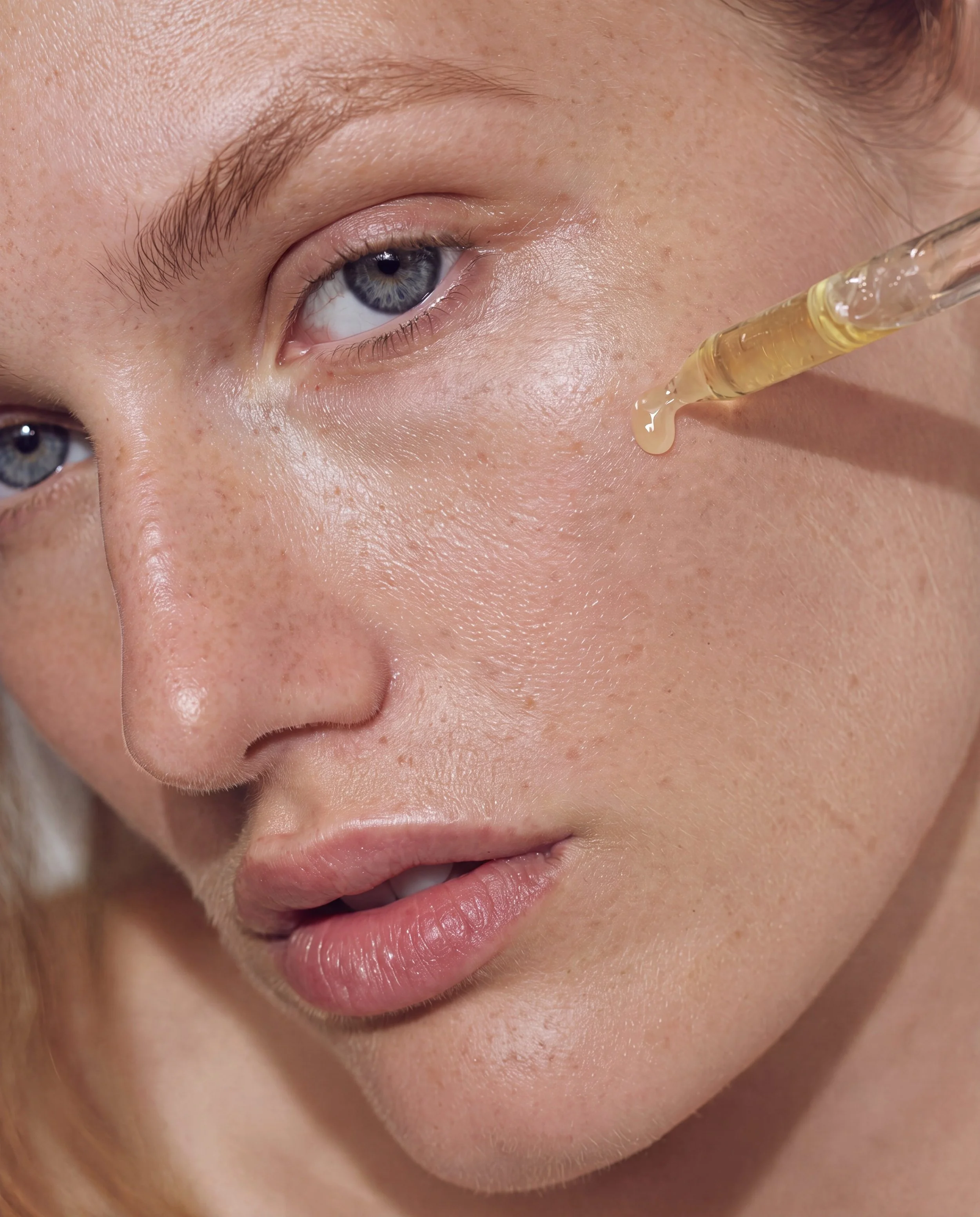 Close-up of a person's face with a dropper applying serum or oil near the eye, showing clear skin, freckles, and light-colored eyebrows.
