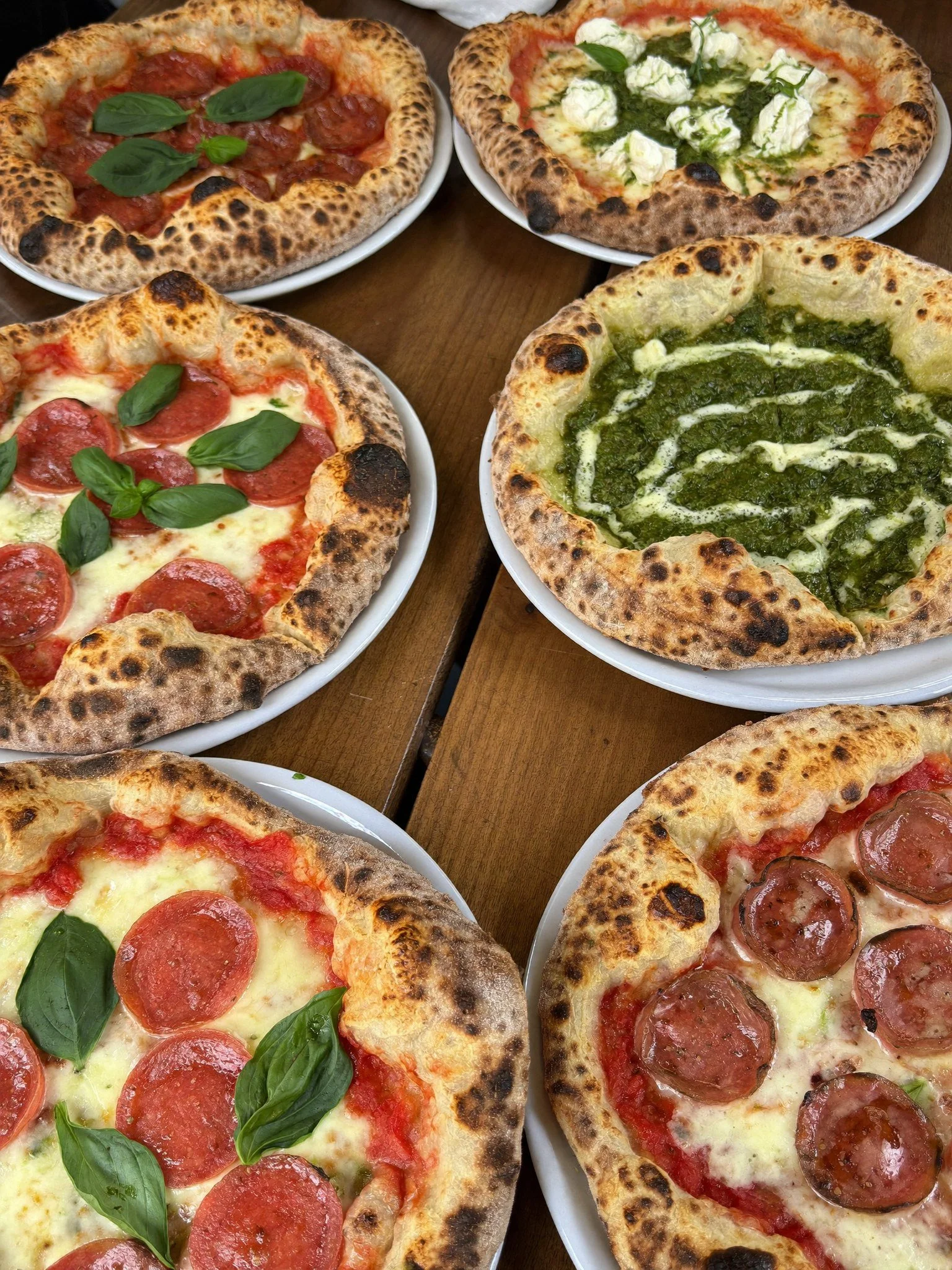 Four pizzas on white plates on a wooden table. Two have pepperoni and basil leaves, one has a green pesto topping with cheese, and another has pepperoni with melted cheese.