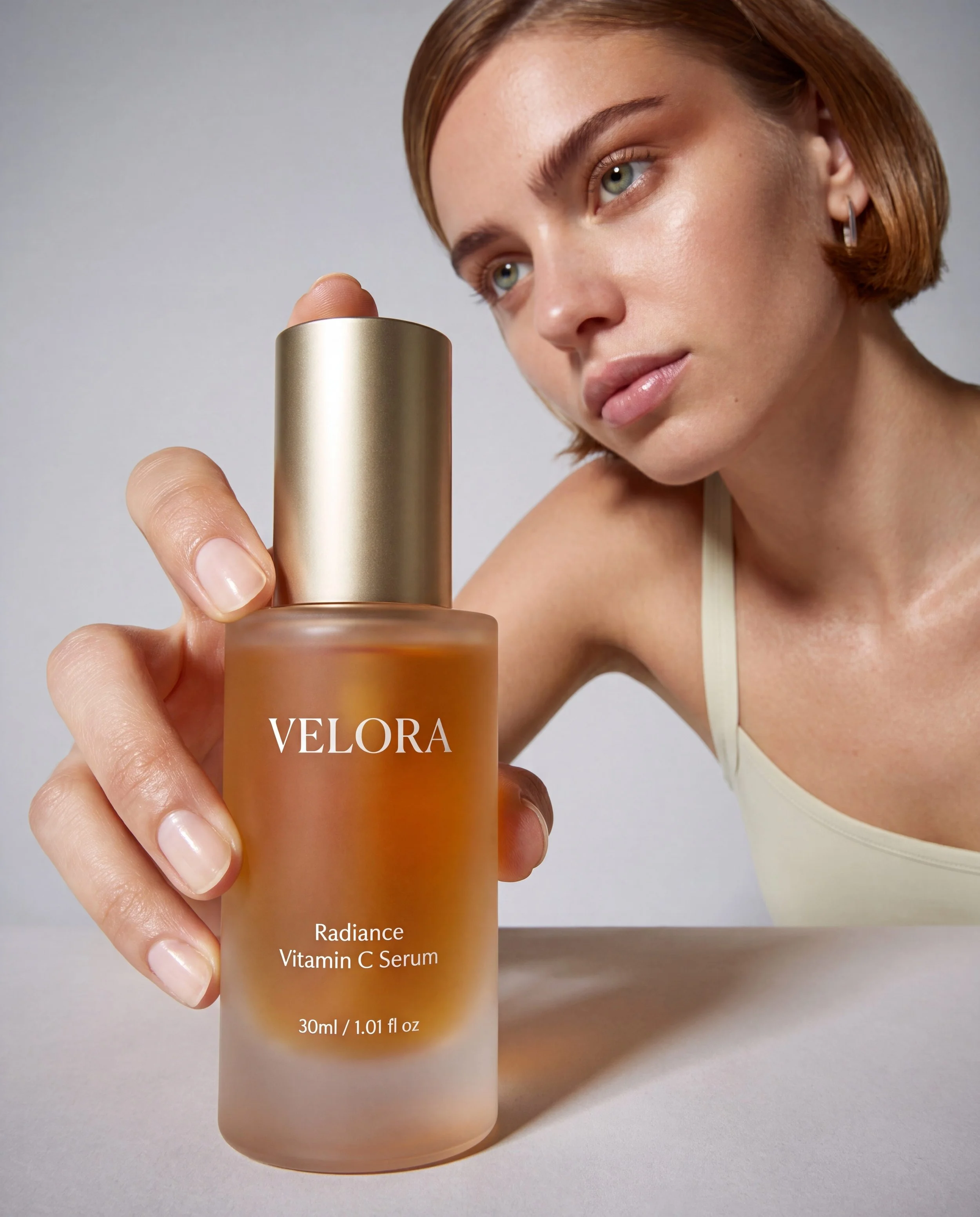 Close-up of a woman with short red hair and fair skin holding a bottle of Velora Radiance Vitamin C Serum. She is wearing a beige tank top, and the background is plain and light-colored.