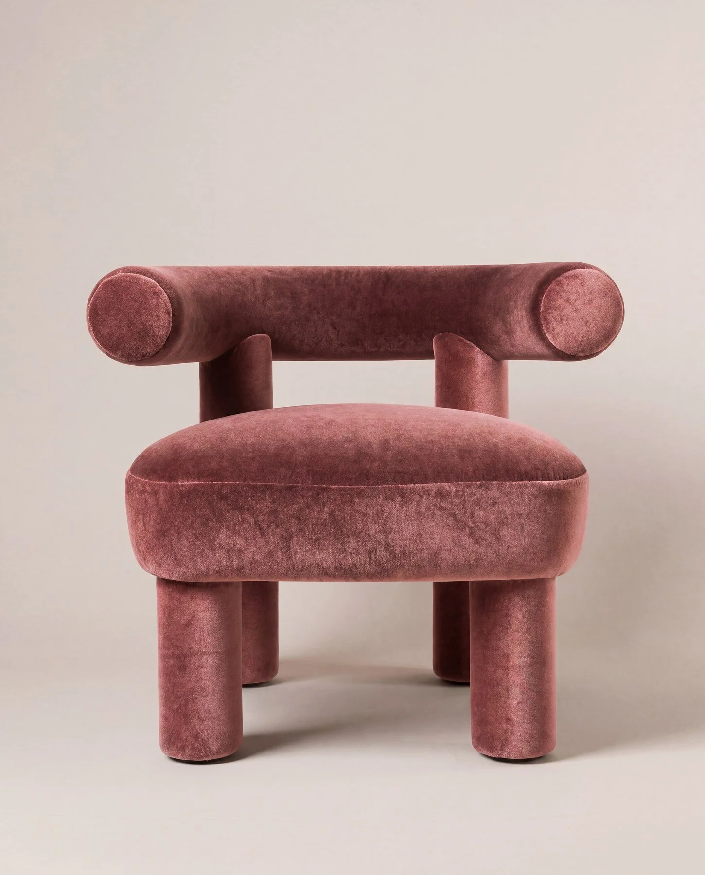 A modern pink velvet chair with rounded armrests and four thick cylindrical legs, set against a plain off-white background.