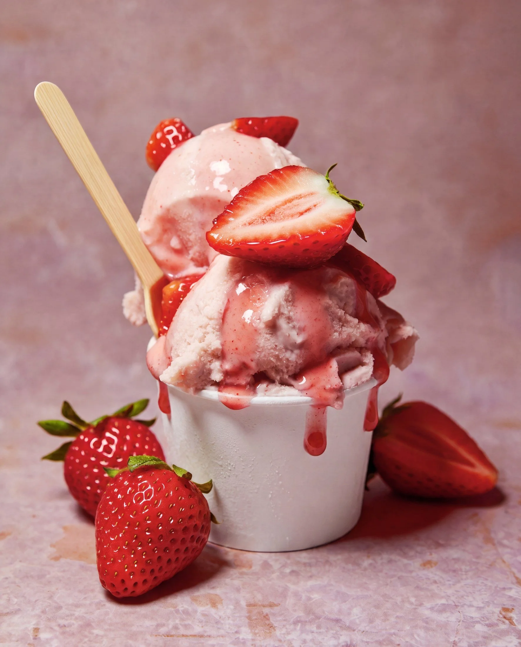 Strawberry ice cream sundae with fresh strawberries on top, drizzled with strawberry syrup, served in a white cup with strawberries on the surface and background.