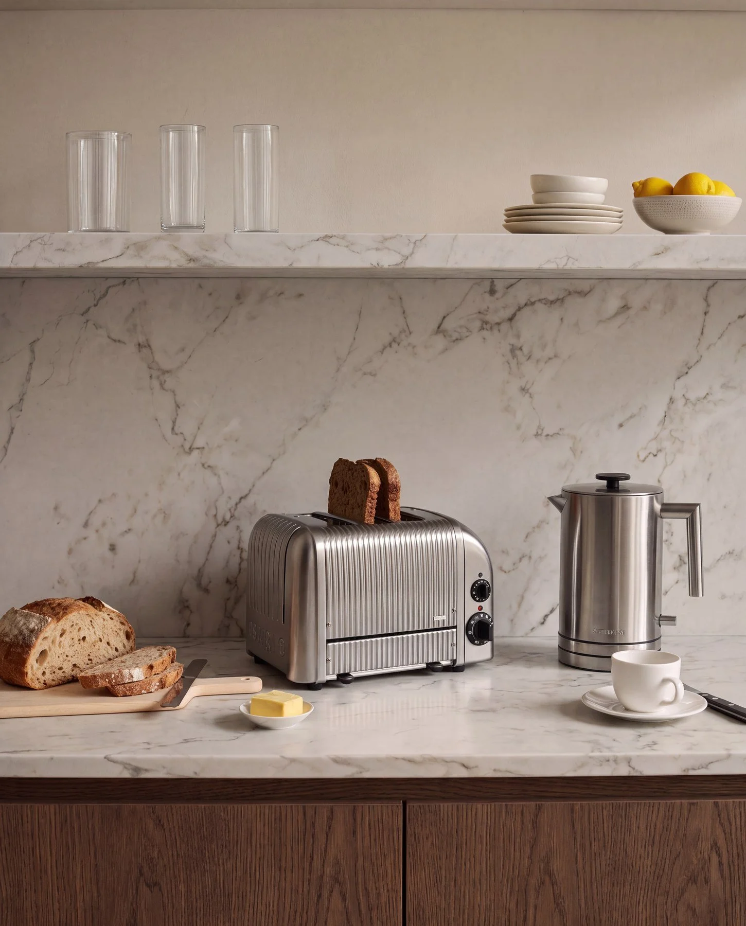 A kitchen countertop with bread, a butter knife, a small dish of butter, a toaster with slices of bread inside, a kettle, a coffee cup on a saucer, and various dishware on shelves above.