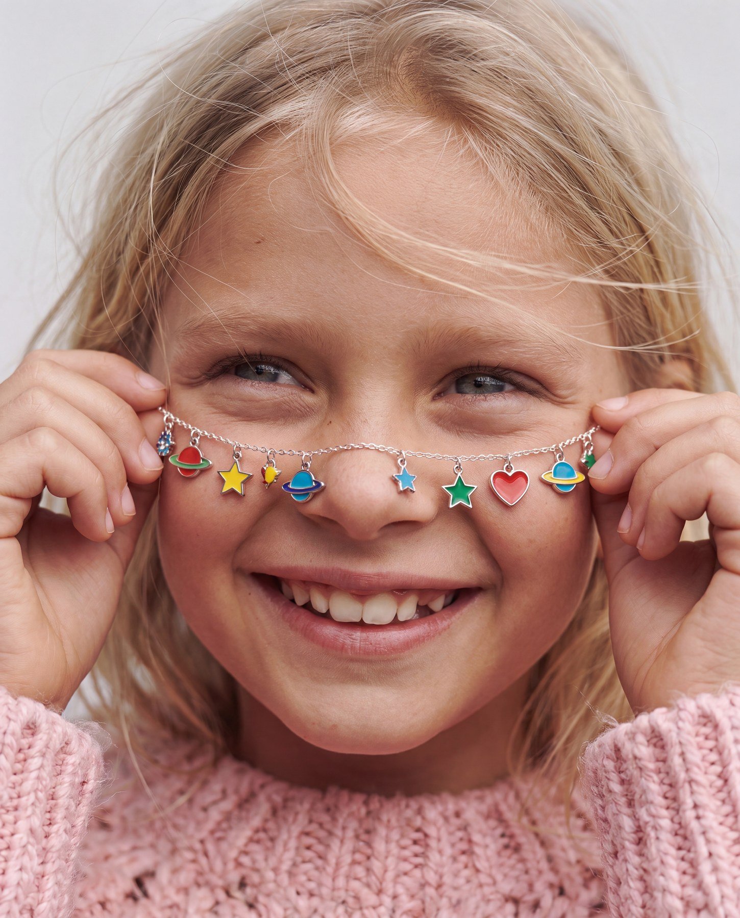 A young girl with blonde hair, smiling and playing with a colorful planet-themed necklace in front of her eyes. She is wearing a pink fluffy sweater.