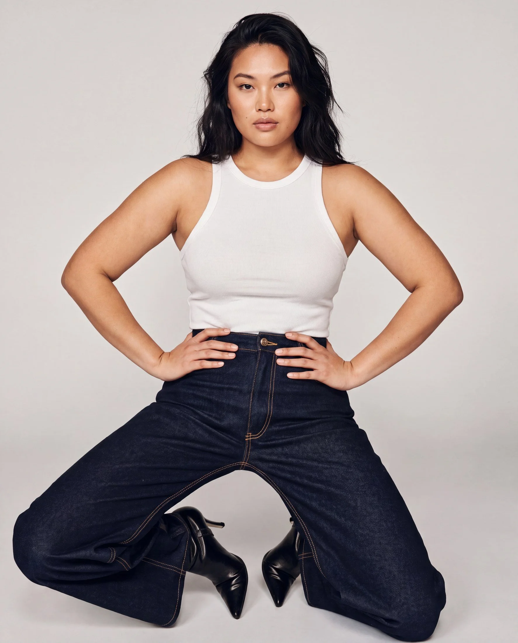 A woman with black wavy hair, wearing a white sleeveless top and dark blue jeans, is posing with her hands on her hips and legs spread apart, against a plain light background.