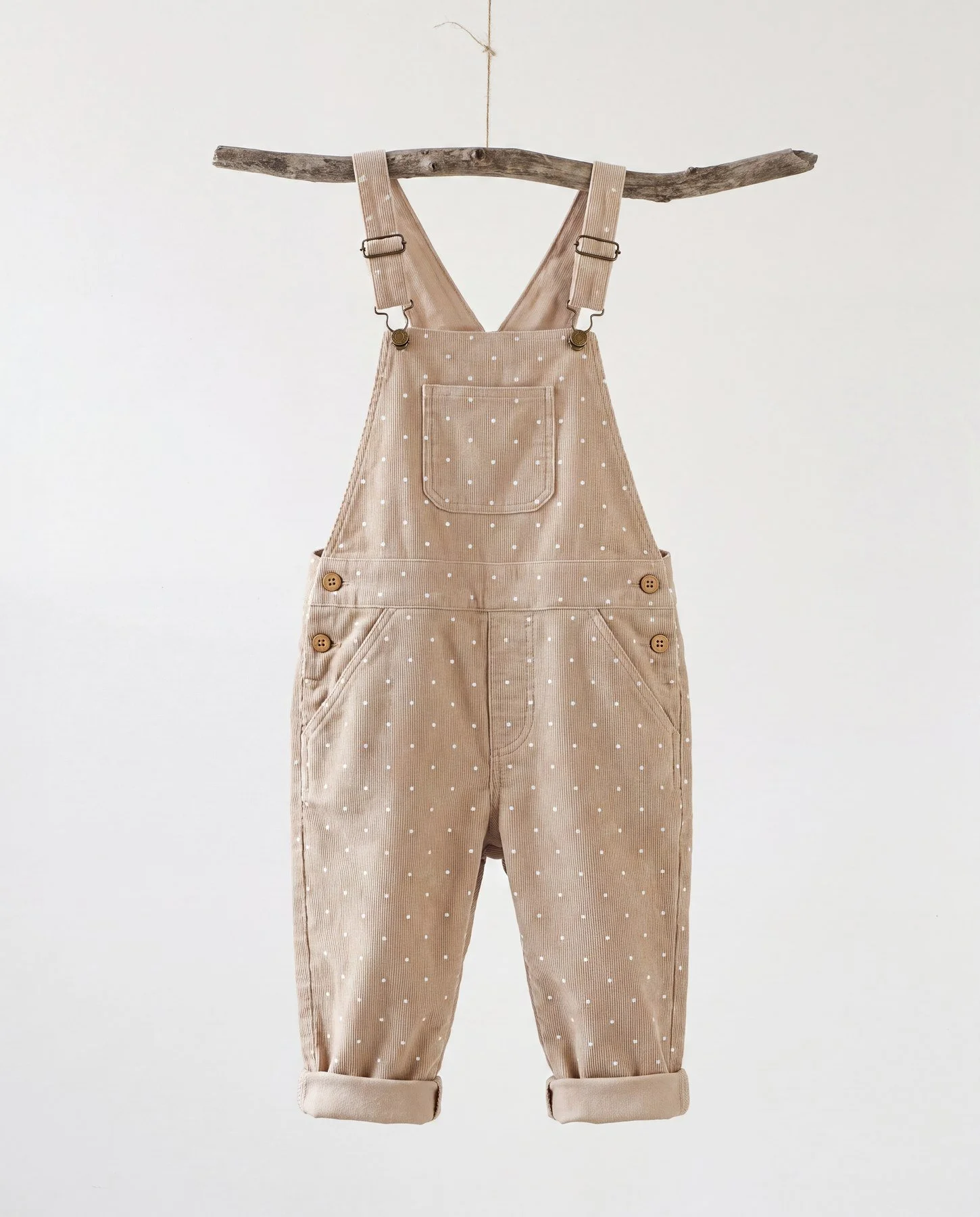 Beige overalls with white polka dots hanging from a wooden branch against a plain white background.