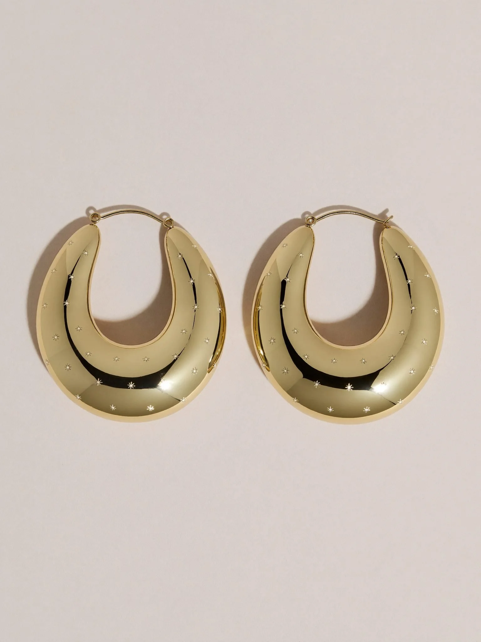 A pair of large, gold hoop earrings with a shiny surface and small star-shaped engravings, placed on a white background.