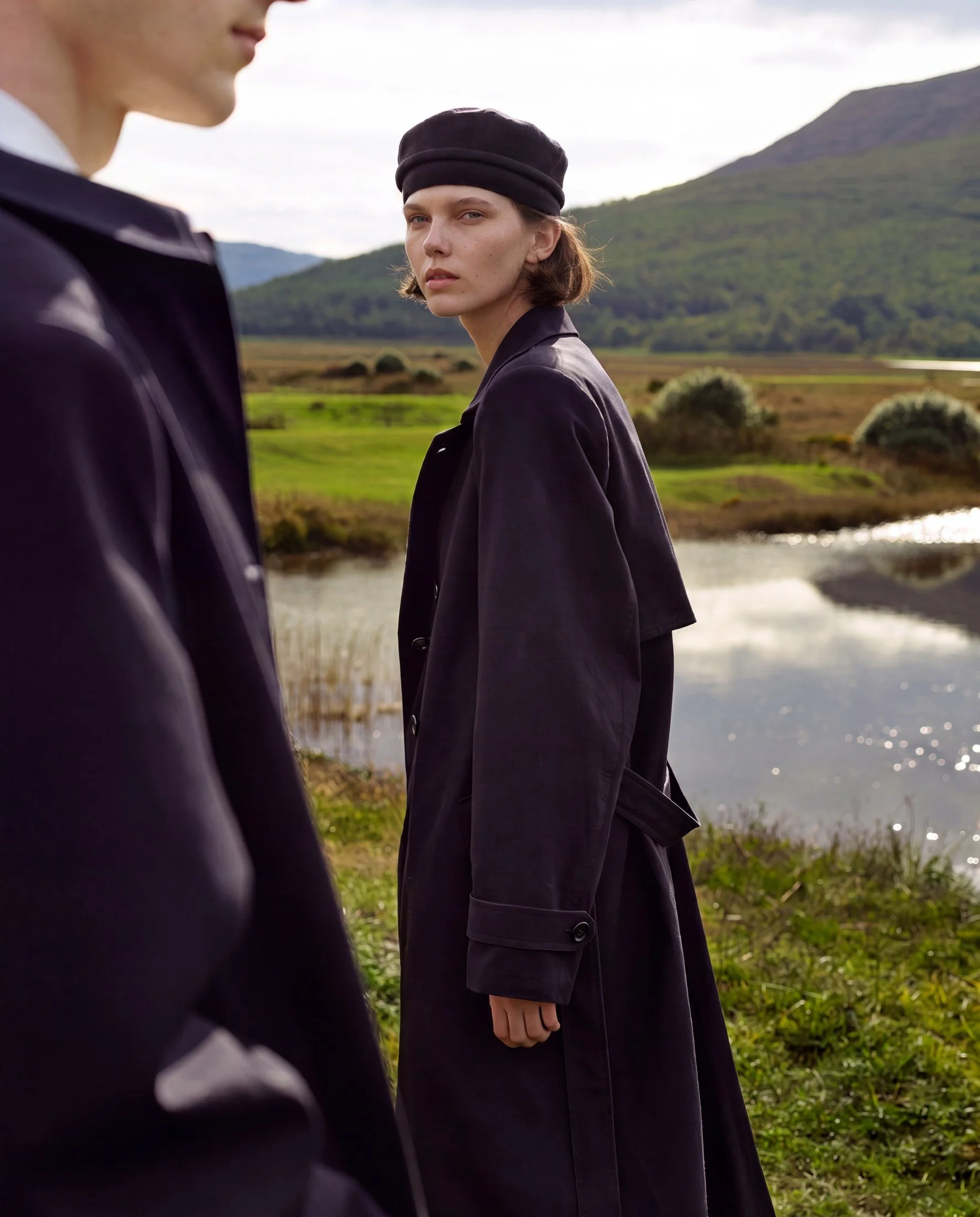 A woman with short brown hair, wearing a black beanie and dark clothing, stands outdoors near a body of water with green hills and mountains in the background, looking at the camera. A part of another person in similar clothing is visible to the left.