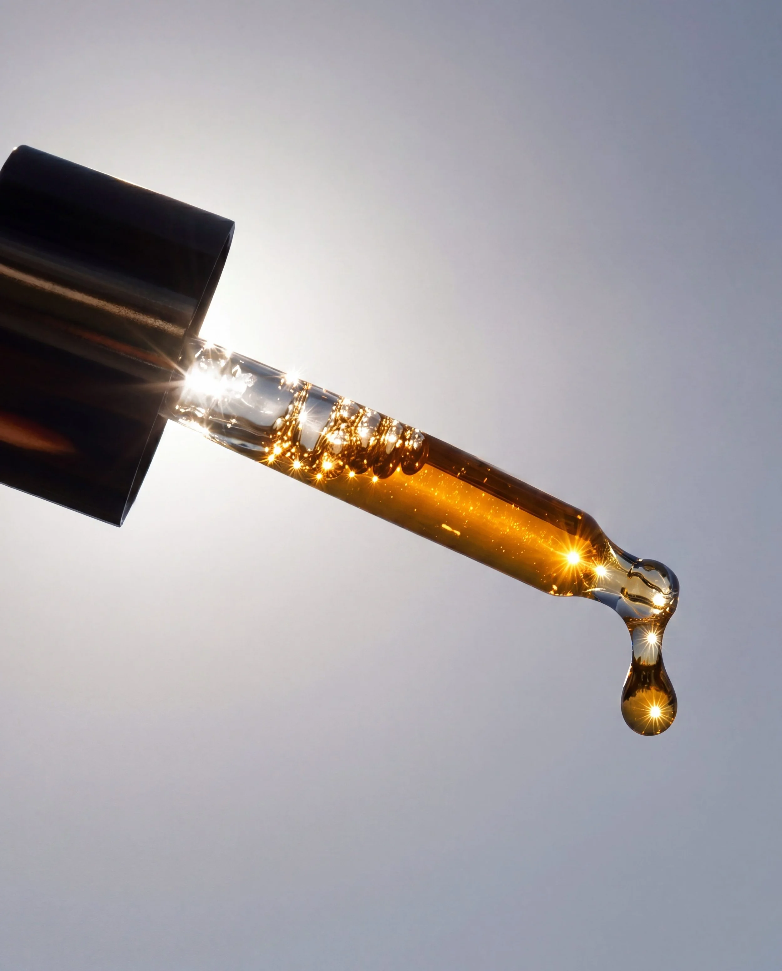 Close-up of a dropper with a golden liquid, shining sunlight reflected on the liquid, against a plain grey background.