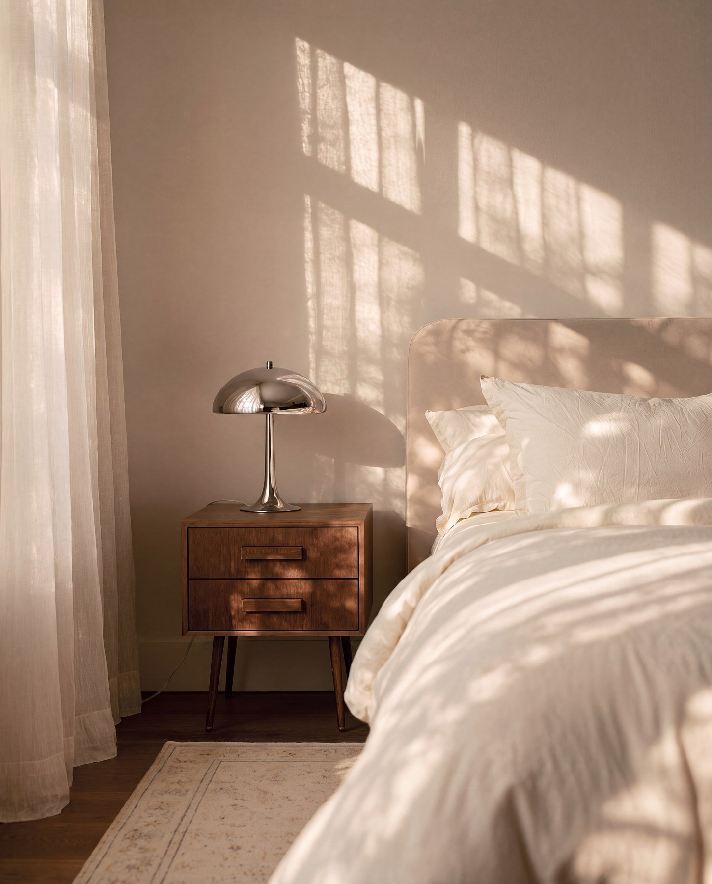 A bedroom corner with a bed, nightstand, and a window casting sunlight and shadows on the wall.