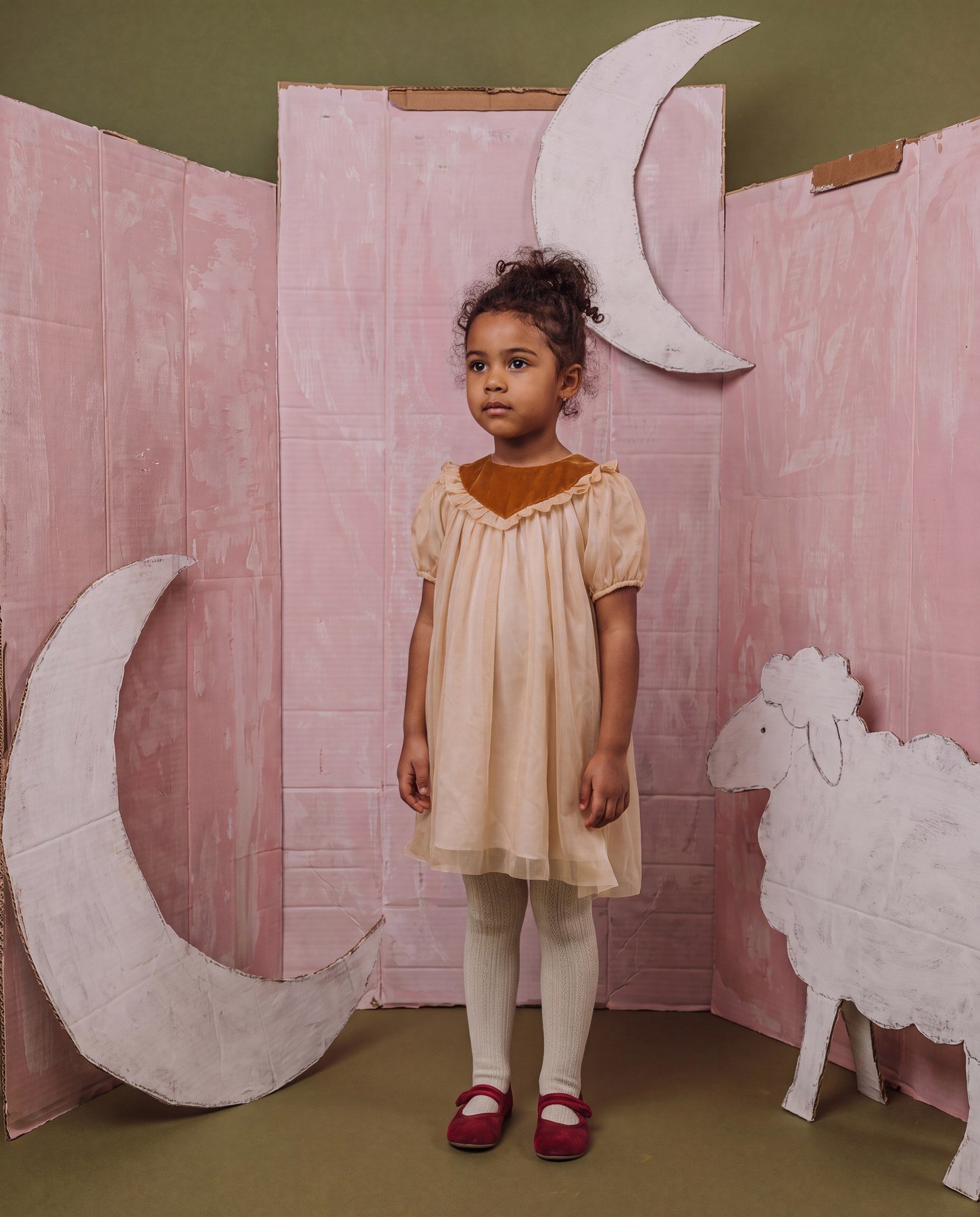 Young girl with curly hair standing in front of a pink cardboard backdrop decorated with white crescent moons and clouds, wearing a beige dress with a brown collar, cream tights, and red shoes.