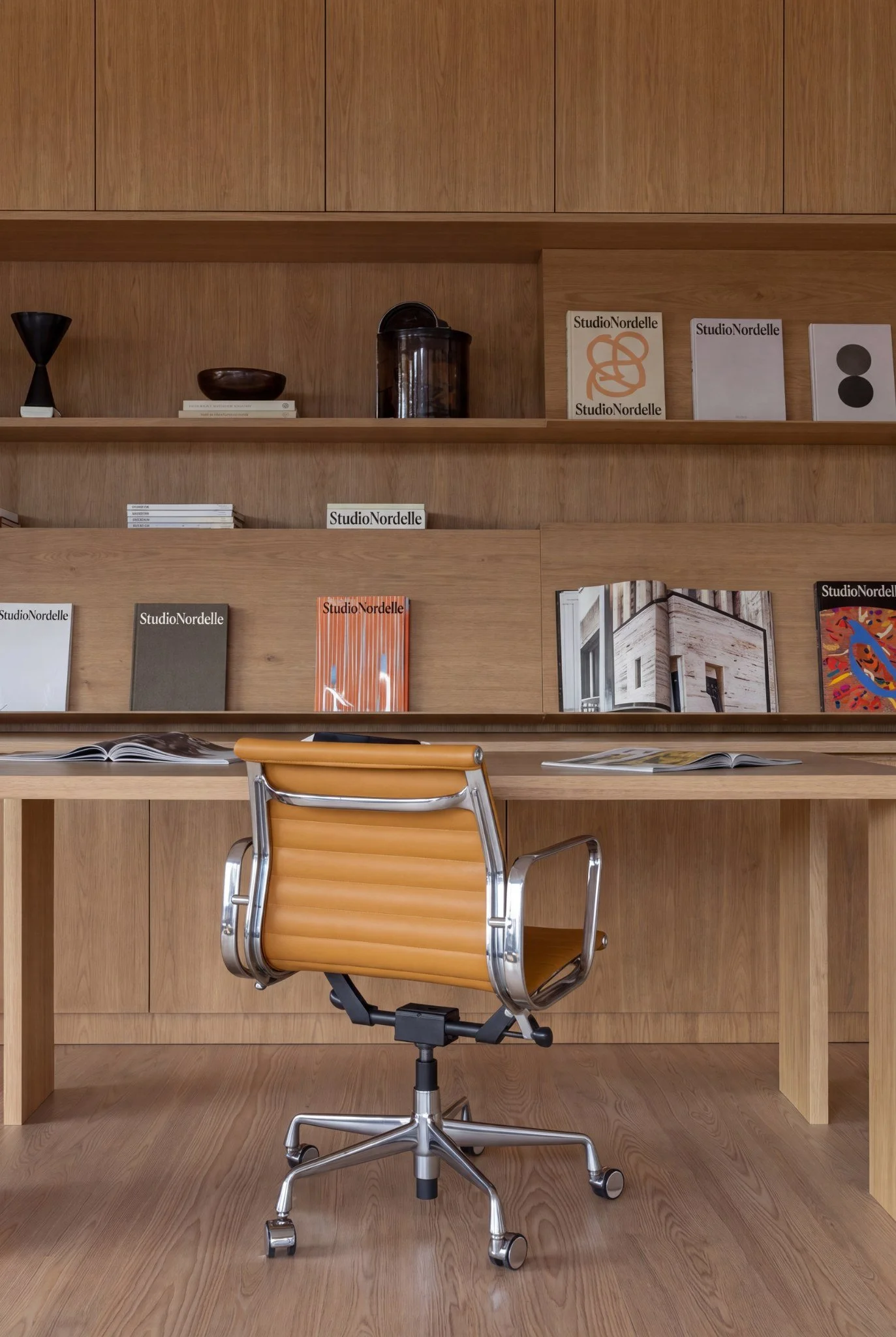 A modern office workspace with a wooden desk, a tan leather office chair, open books and magazines, a bookshelf with various books and decorative items, and a wood-paneled wall with artwork and decorative objects.
