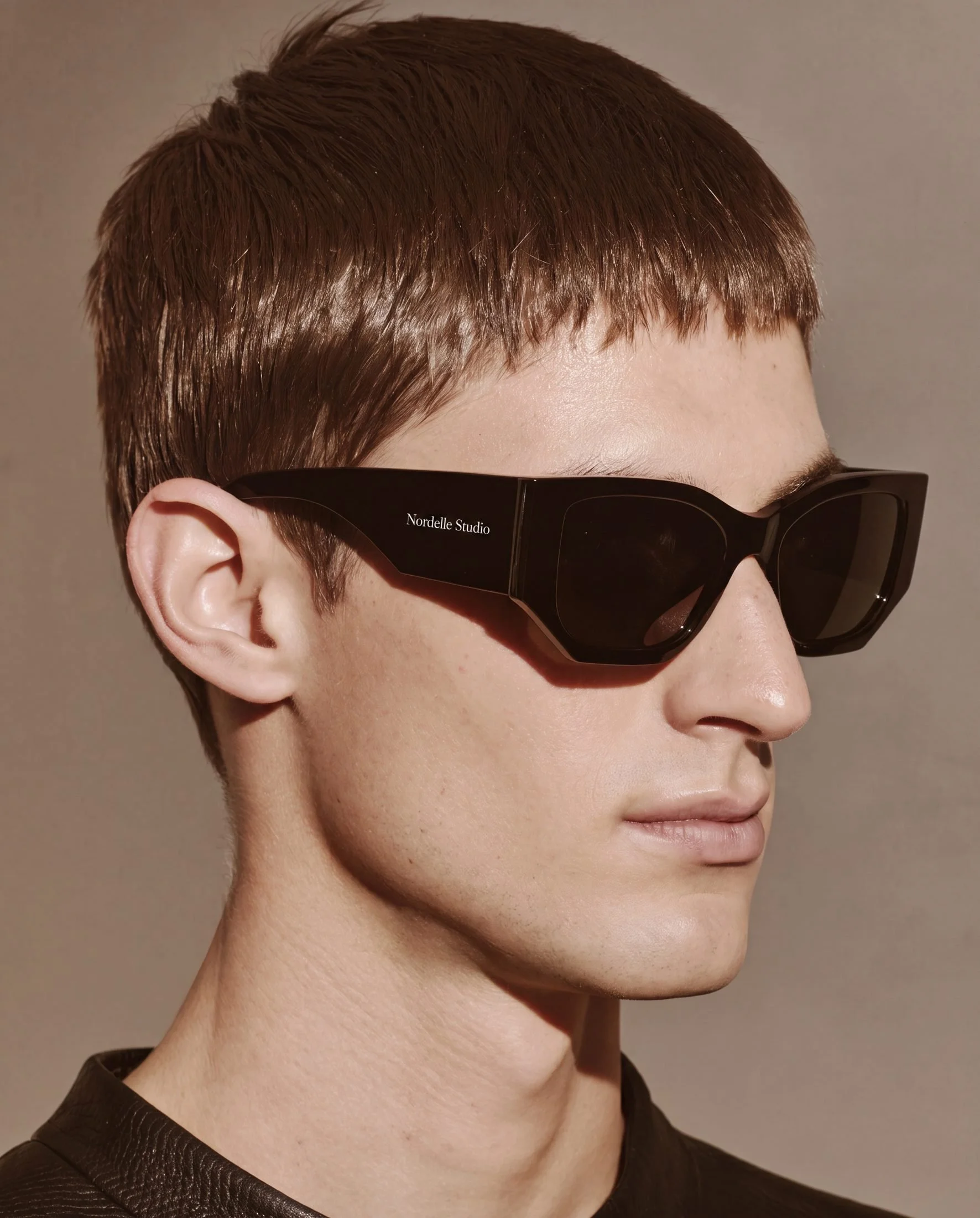 Close-up of a person wearing black sunglasses with brown hair and a short haircut, dressed in a black textured top, against a plain background.
