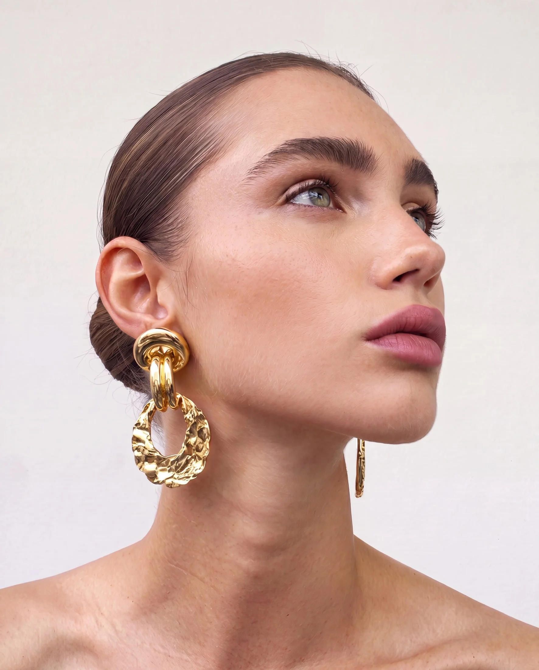 Close-up of a woman's face showing her makeup, hair, and gold hoop earrings, with a neutral background.