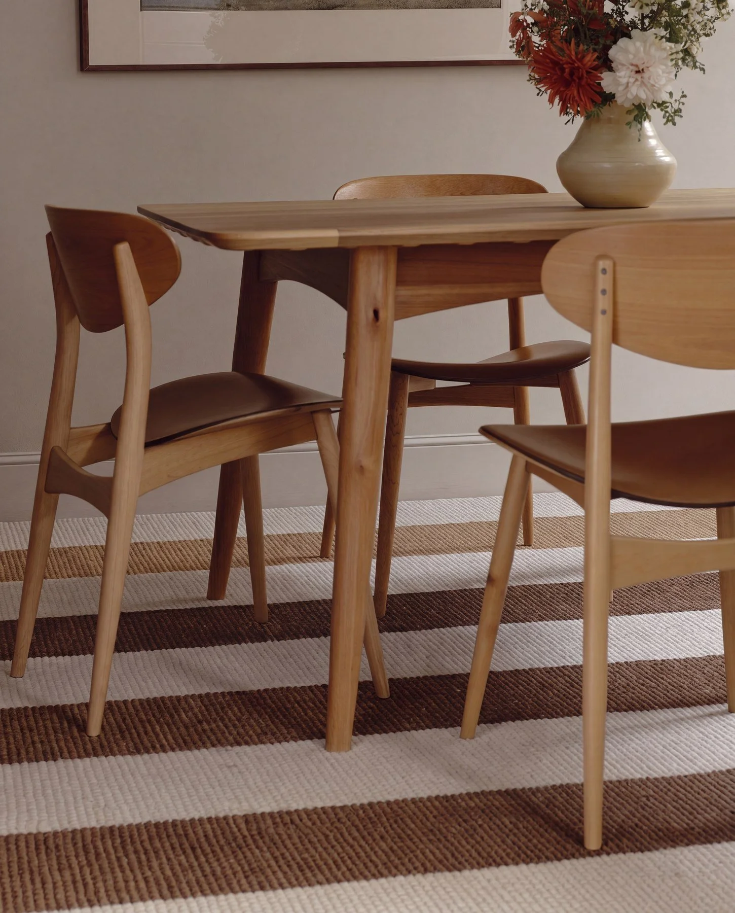 A wooden dining table with four matching chairs. There is a vase with flowers on the table and a framed picture hanging on the wall in the background. A striped rug is underneath the table.
