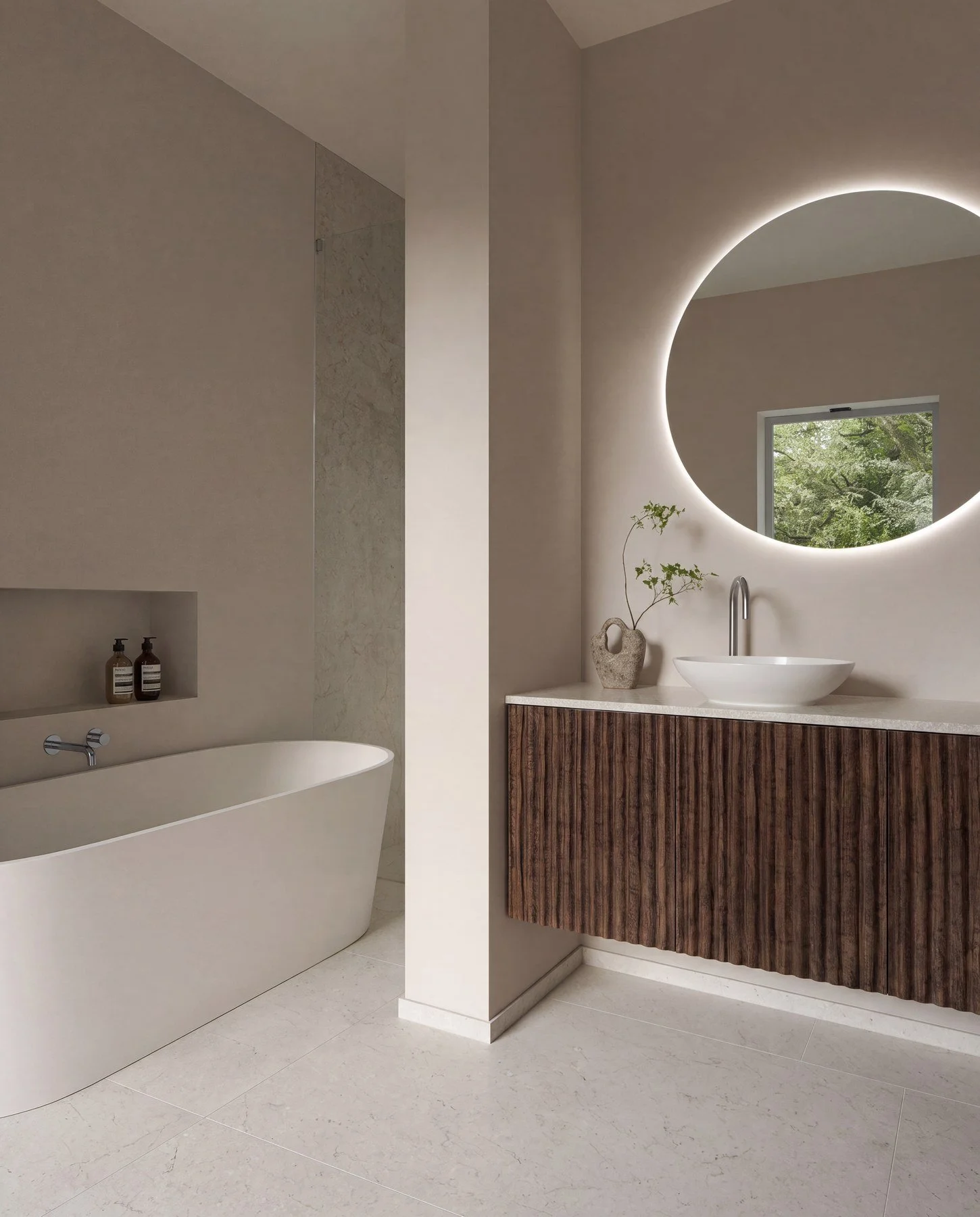 Modern bathroom with a white freestanding bathtub and a wooden vanity with a circular mirror illuminated by LED backlighting, a small tonked vase with green branches, and a window showing green trees outside.