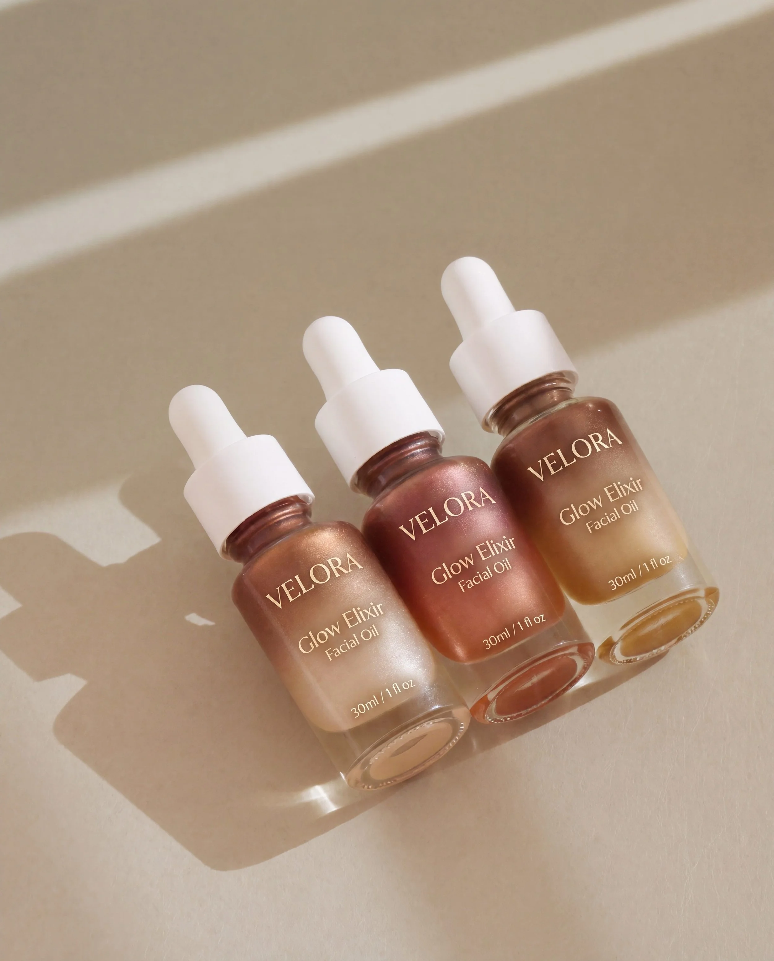 Three bottles of Velora Glow Elixir Facial Oil arranged in a diagonal row on a neutral surface. The bottles have white dropper caps and labels with the product name and description.