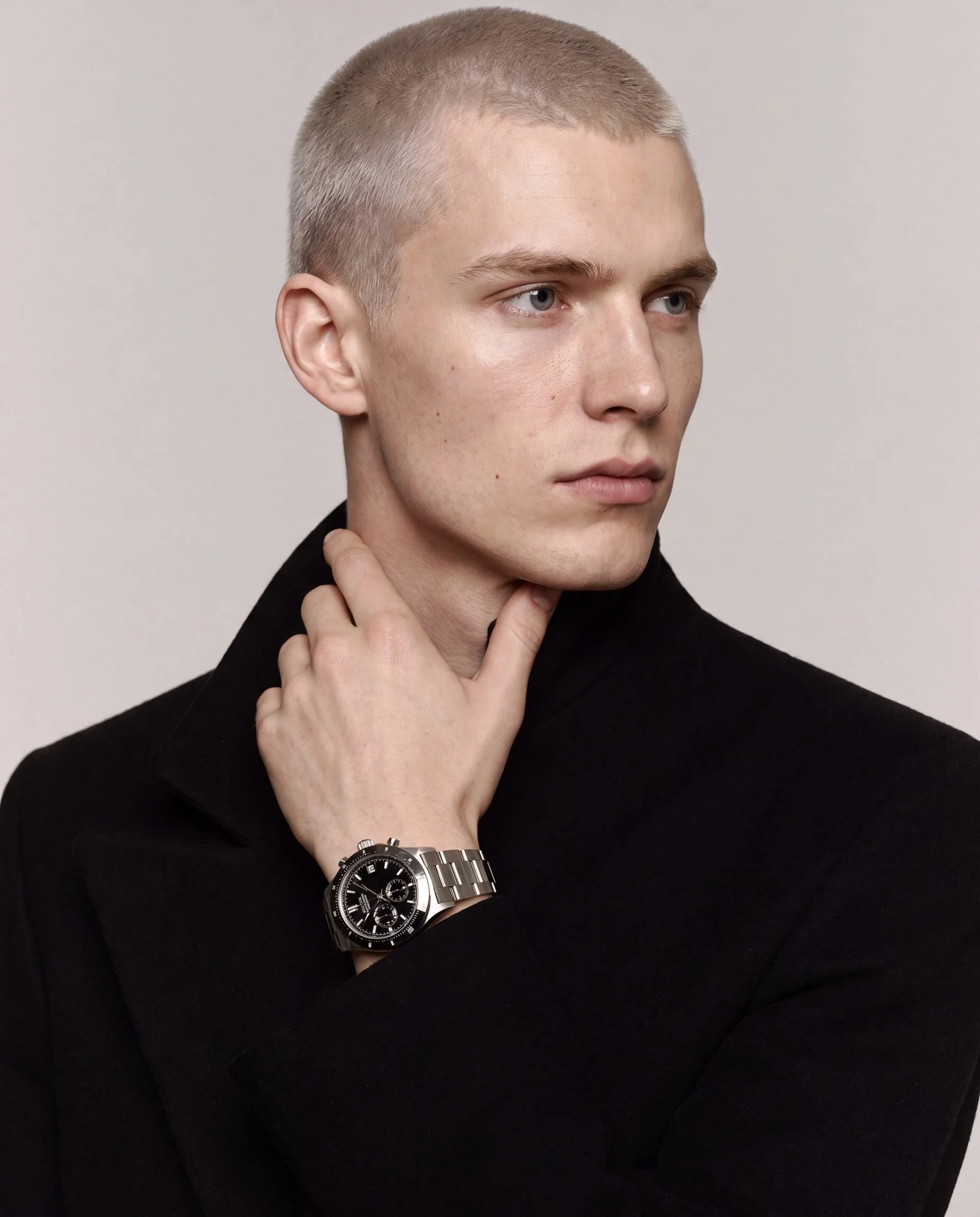 A young man with short, light-colored hair, blue eyes, and fair skin, wearing a black turtleneck and a large wristwatch, touching his neck with his right hand, against a plain grey background.