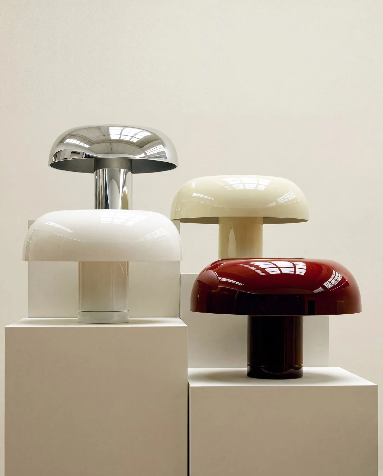 Display of four modern, glossy mushroom-shaped stools in different colors: silver, white, red, and beige, arranged on white pedestals against a plain white wall.