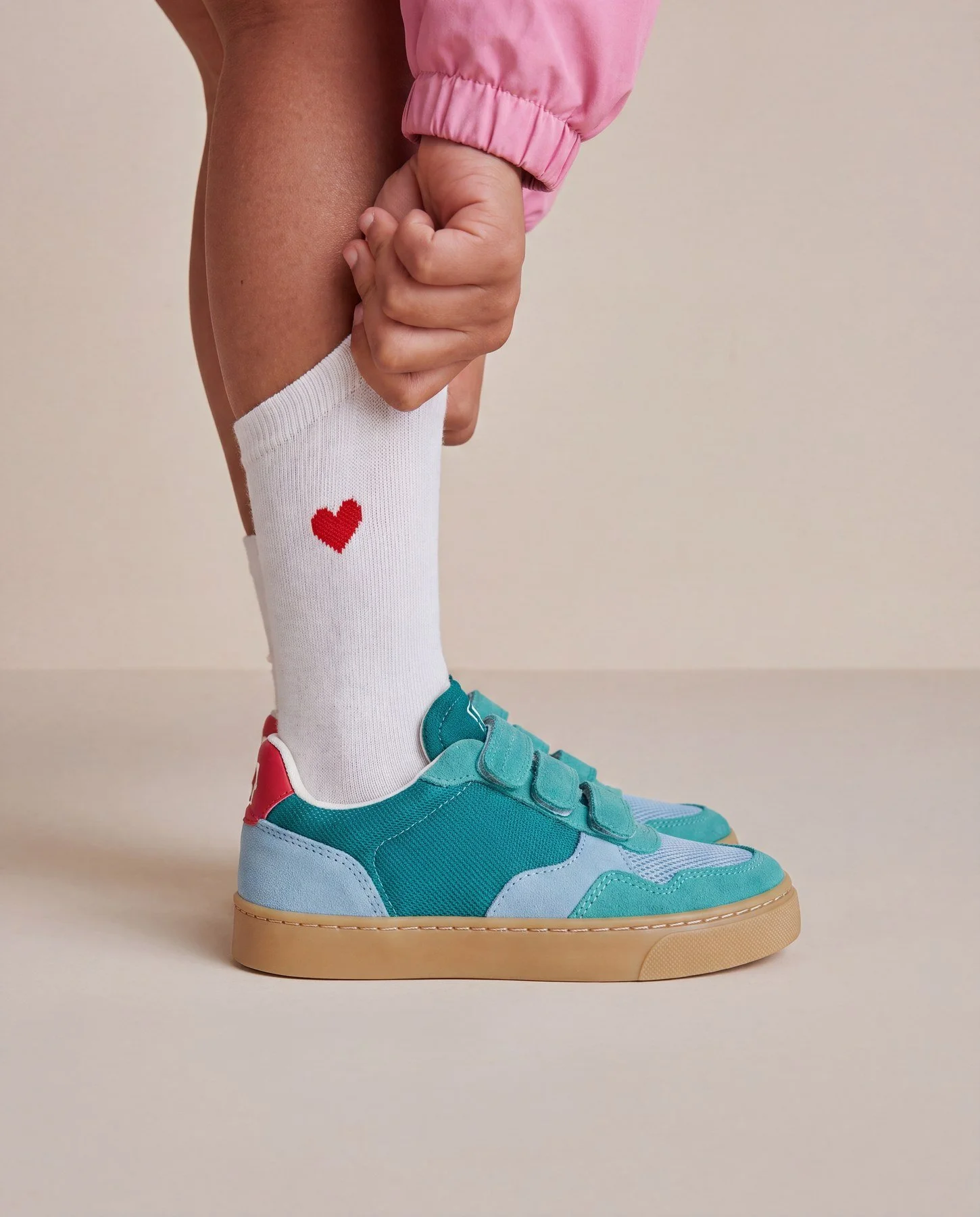 A person wearing white socks with red heart design, blue sneakers, pink jacket, and tan sneakers, with their hand clutching their knee.