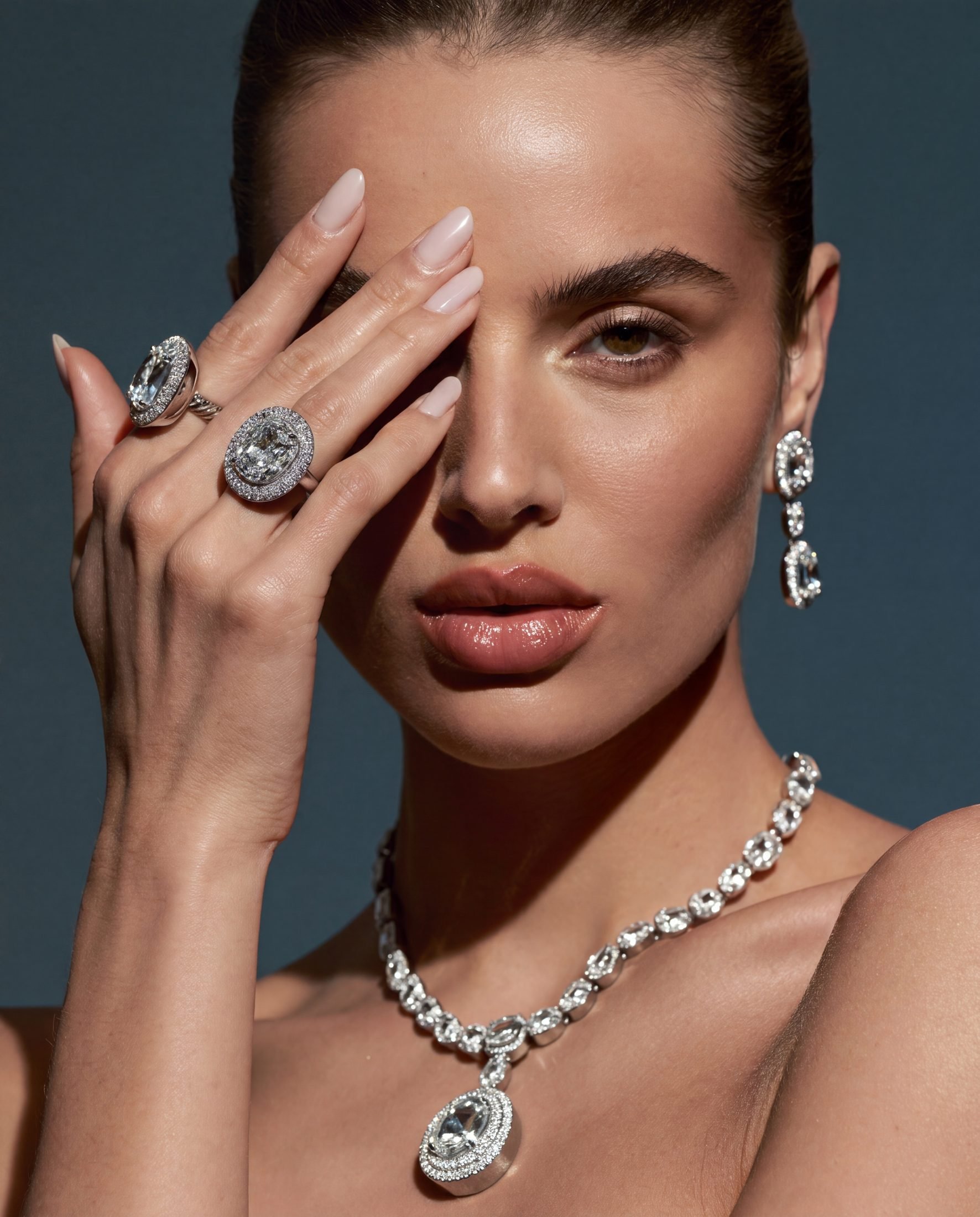 Close-up of a woman wearing diamond jewelry, including rings, earrings, a necklace, and a large pendant. She is holding her hand with rings up to her face, covering one eye.