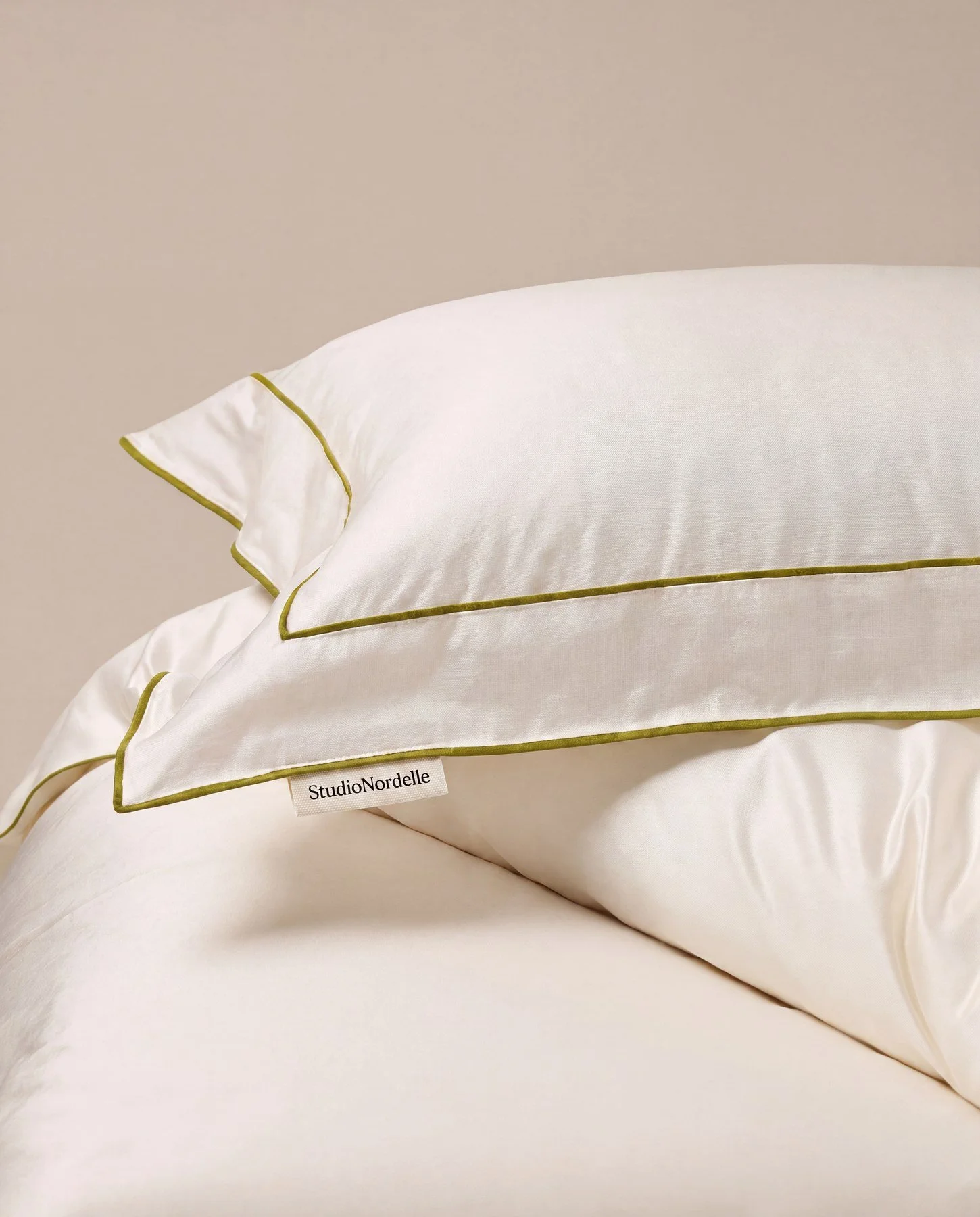 Set of white satin pillows with green piping, stacked on top of each other, with a label reading 'StudioNordelle' on the lower pillow.