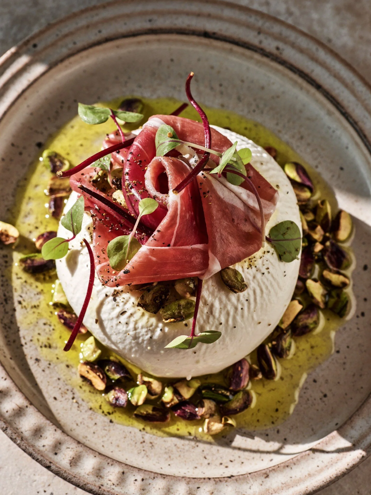 A dish featuring a round white cheese topped with thin slices of prosciutto, microgreens, and crushed pistachios, drizzled with olive oil on a speckled ceramic plate.