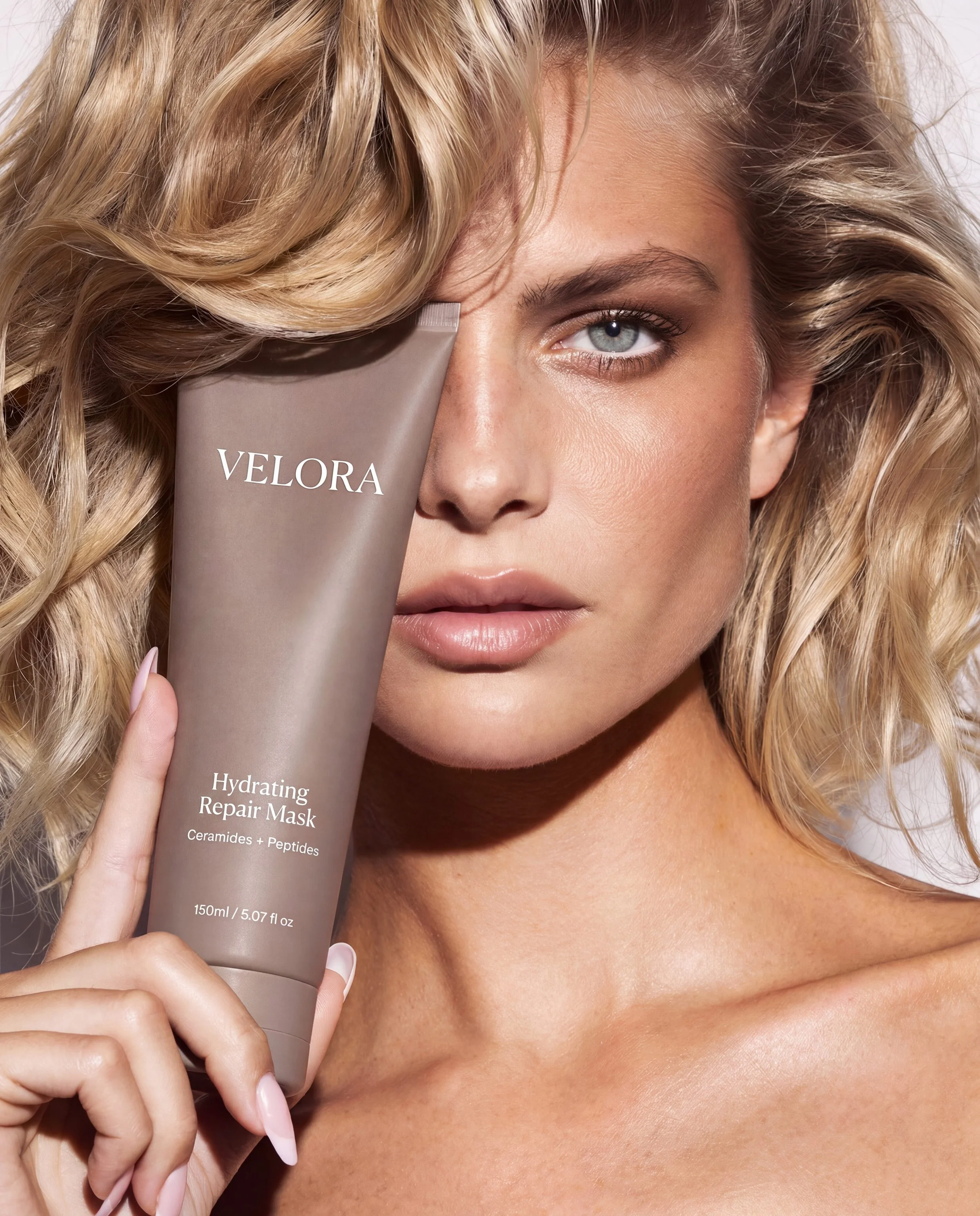 A woman with blonde hair and blue eyes holding a tube of Velora Hydrating Repair Mask in front of her face. The focus is on her face and the product.