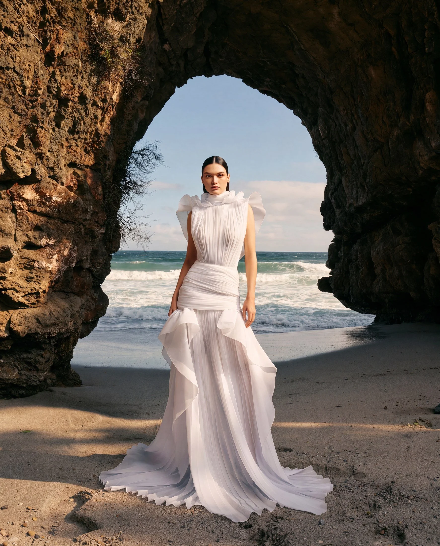 A woman in a flowing white dress standing on a beach, framed by a large rock archway with the ocean and a partly cloudy sky in the background.