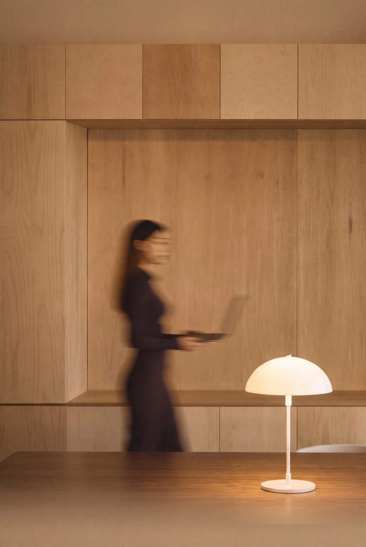 A woman in black walking past a wall, holding a tablet, with a white table lamp on a wooden surface in the foreground.