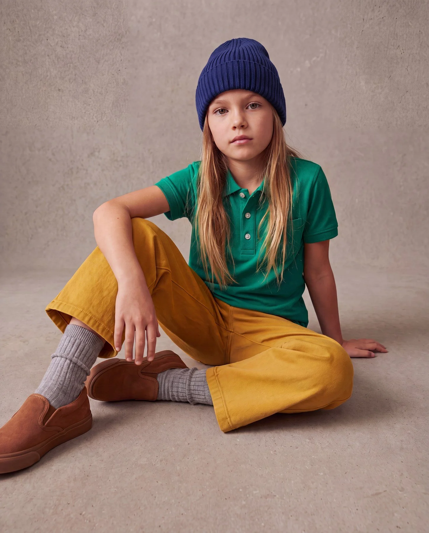 A young girl sitting on the floor, wearing a blue knit beanie, a green polo shirt, mustard yellow pants, gray socks, and brown shoes, against a plain beige background.