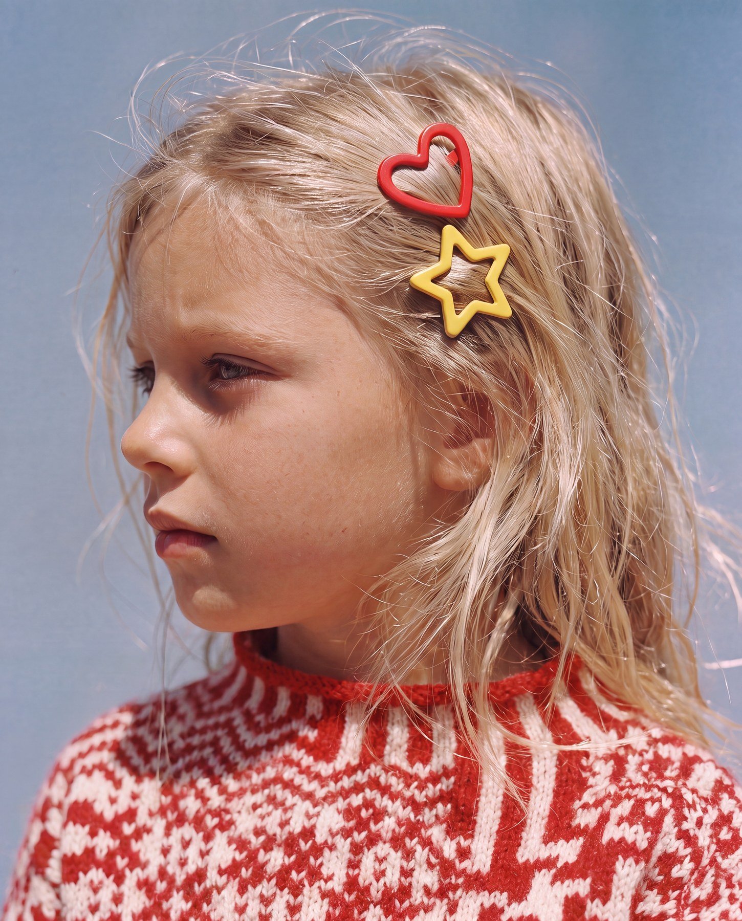 A young girl with wet blonde hair, wearing red and white patterned sweater, has two colorful hair clips in the shape of a heart and a star in her hair, against a plain background.