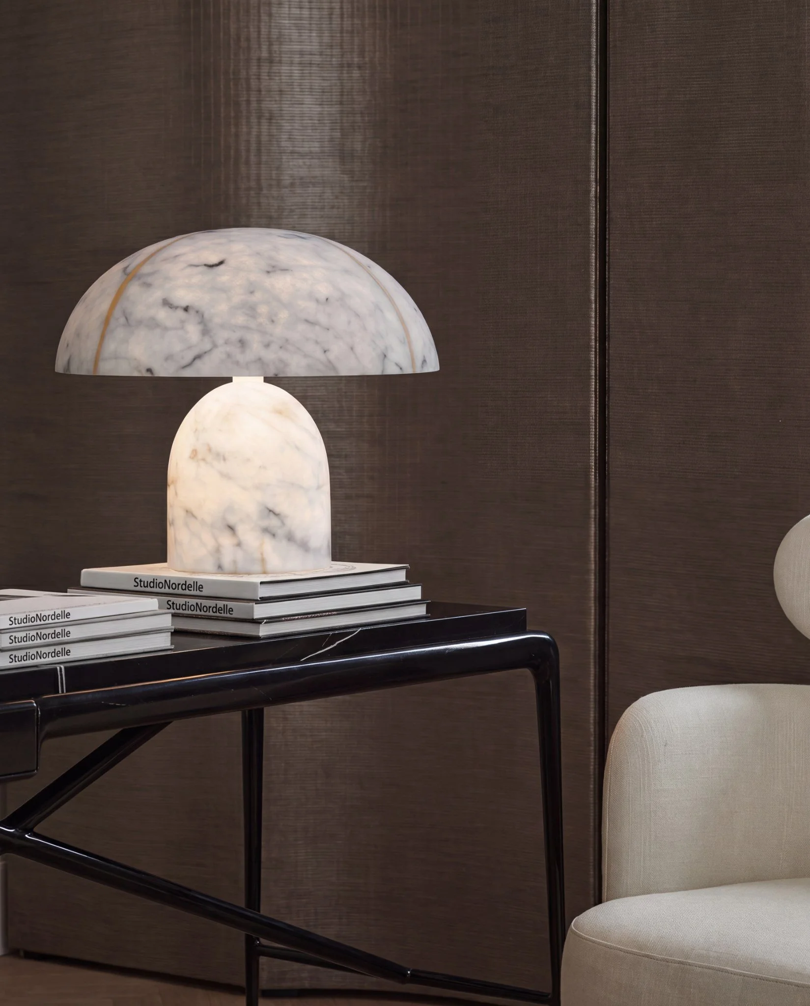 A modern table lamp with a marble base and a dome-shaped shade on top, placed on a table with books labeled 'Studio Nordelle' beneath it. The background features a brown textured wall and part of a beige chair.